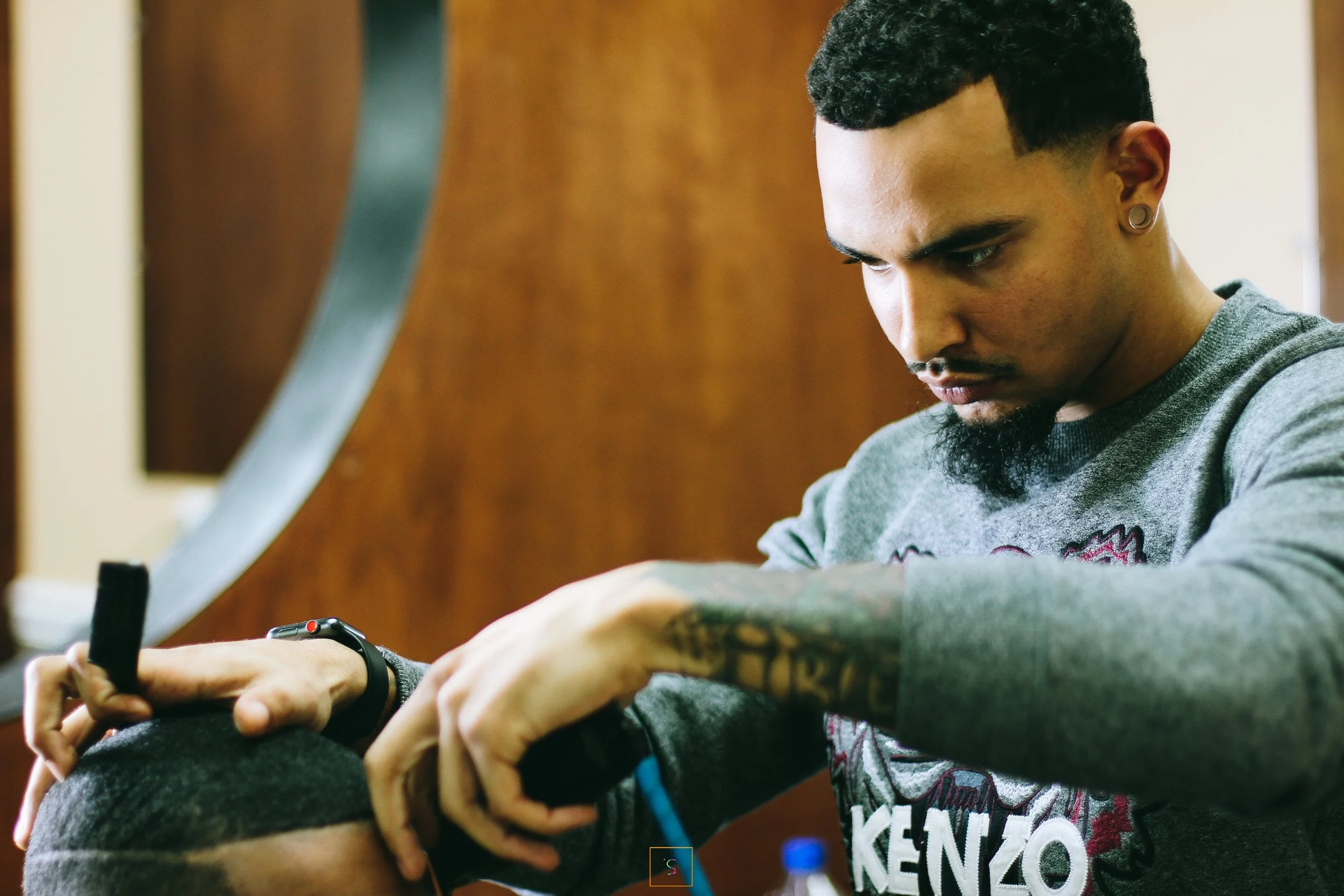 A man with tattoos and earrings is looking down while holding a black object, wearing a gray sweatshirt with multicolored details, in an indoor setting with wooden walls.