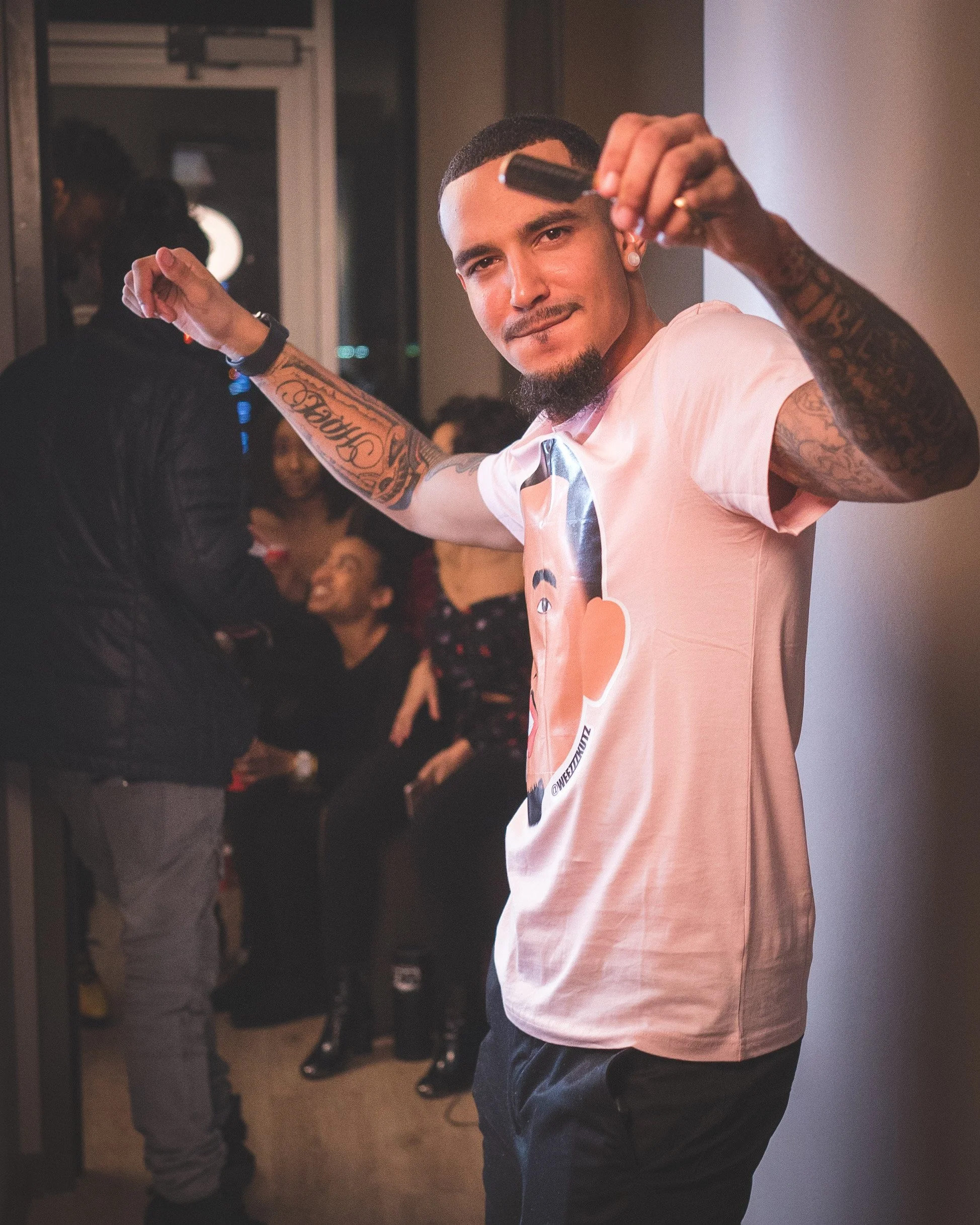 A man with tattoos, a goatee, and a short haircut, holding a razor in front of him, posing at a social gathering indoors with people in the background.