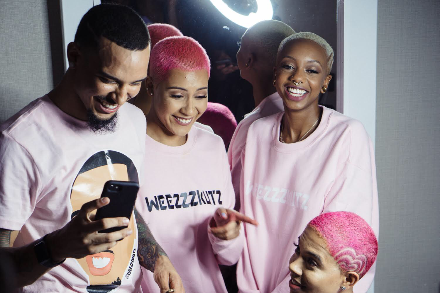 A group of five smiling people with pink hair, wearing matching pink shirts, taking a selfie together indoors.