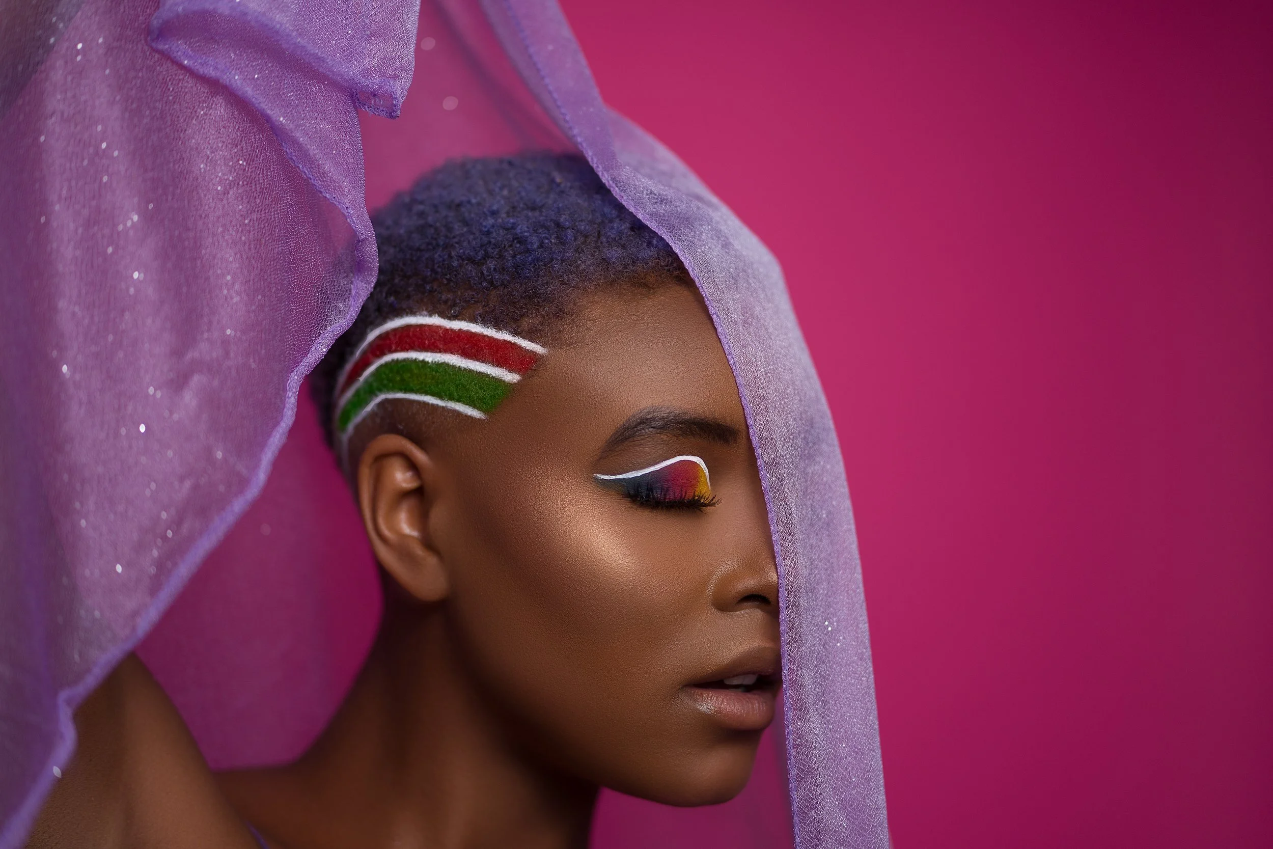 Close-up of a woman with colorful rainbow-themed eye makeup and a purple sheer veil, with sides of her head decorated with red, white, and green stripes.