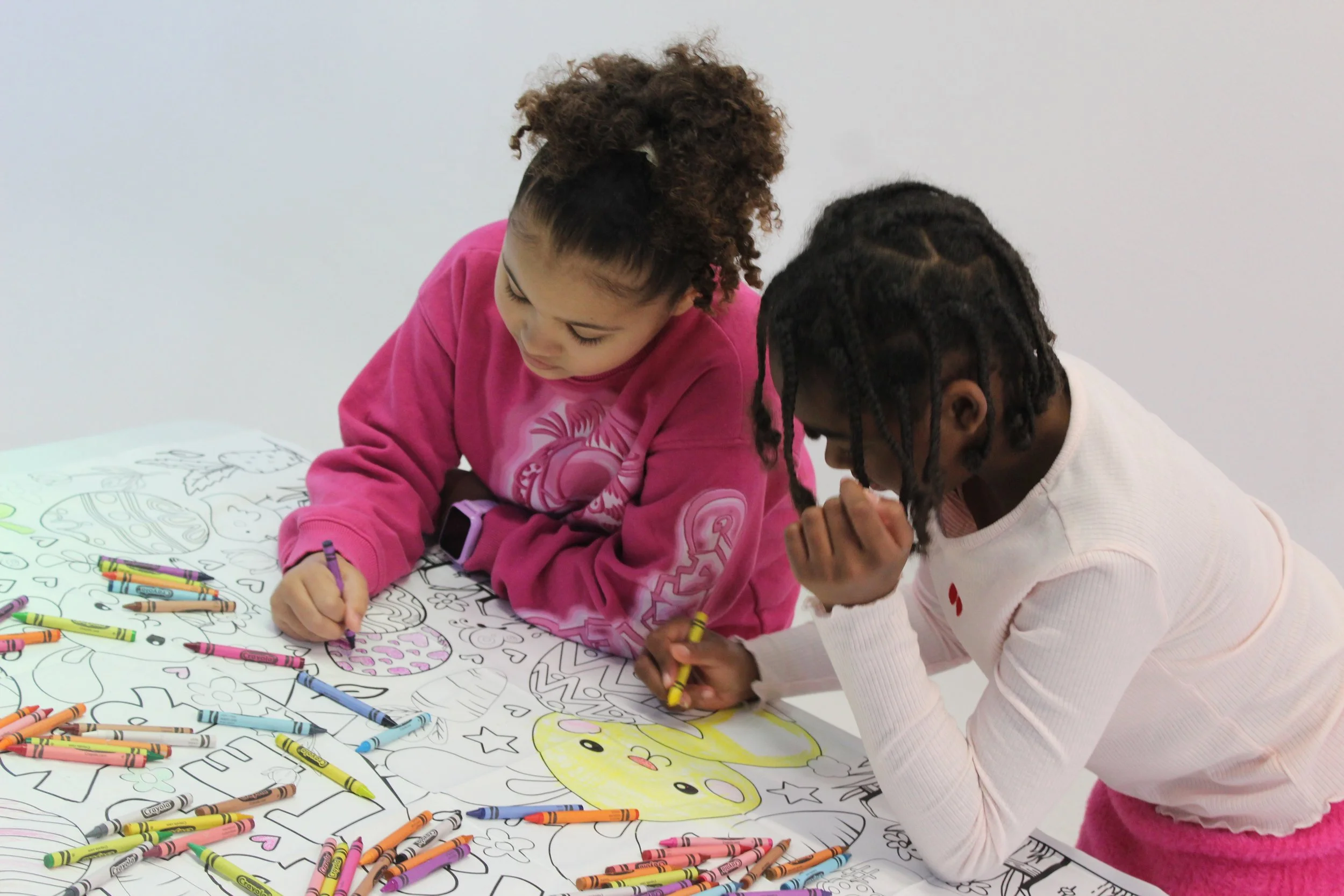Two young girls are coloring a large picture made with outlines on a giant sheet of paper, with many crayons scattered around.