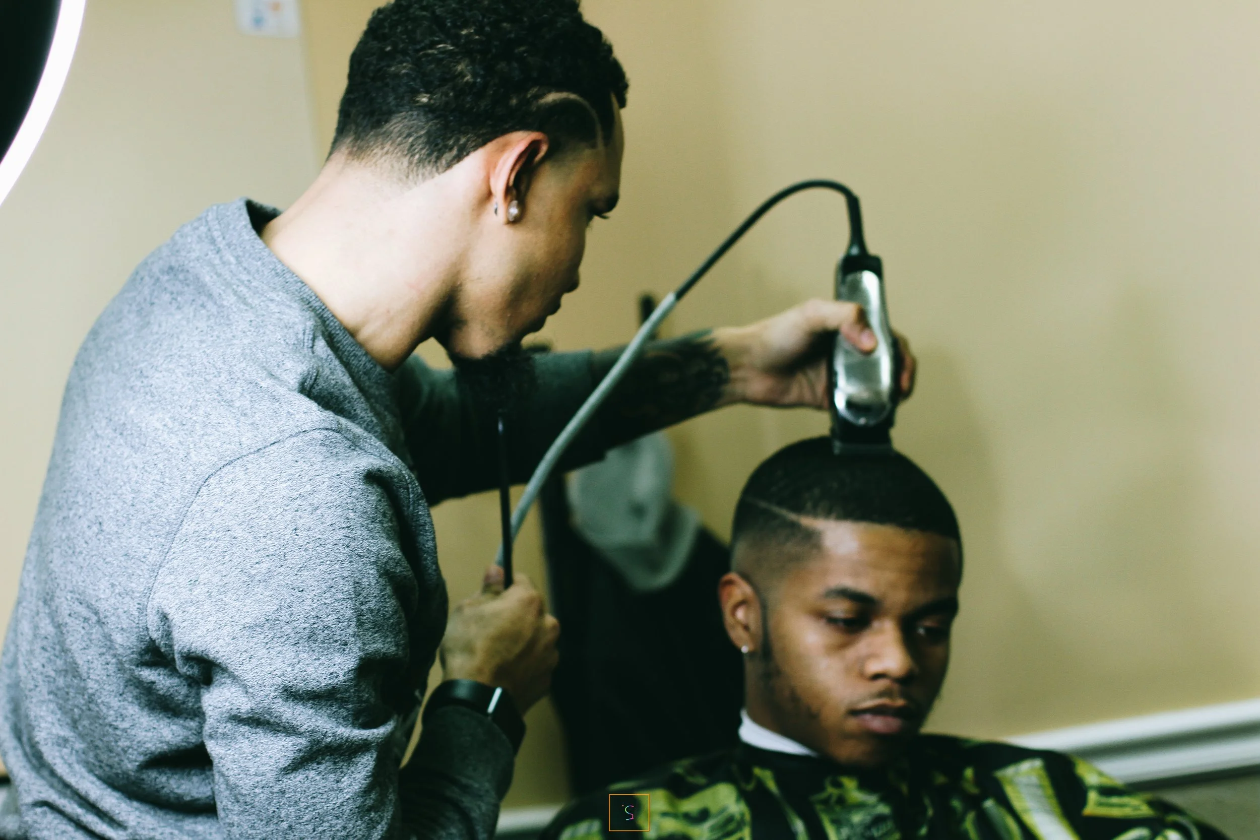 Barber trims a young man's hair using clippers in a barbershop, with the young man seated and wearing a green and black patterned cape.