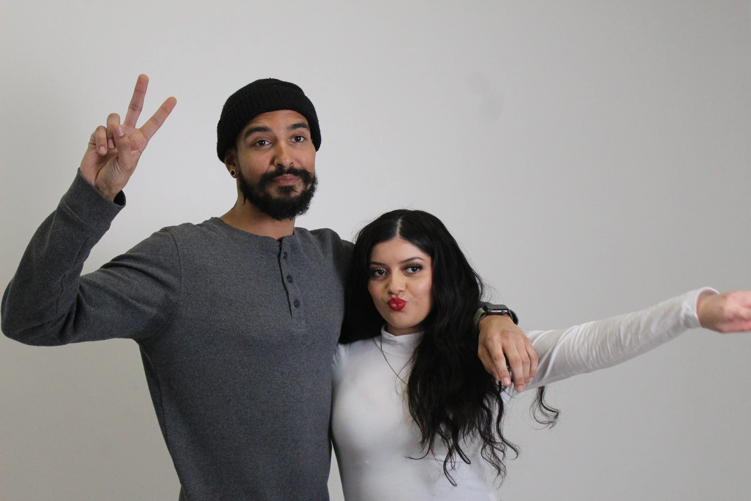 A man and woman posing together against a plain grey wall, making peace signs and playful gestures.