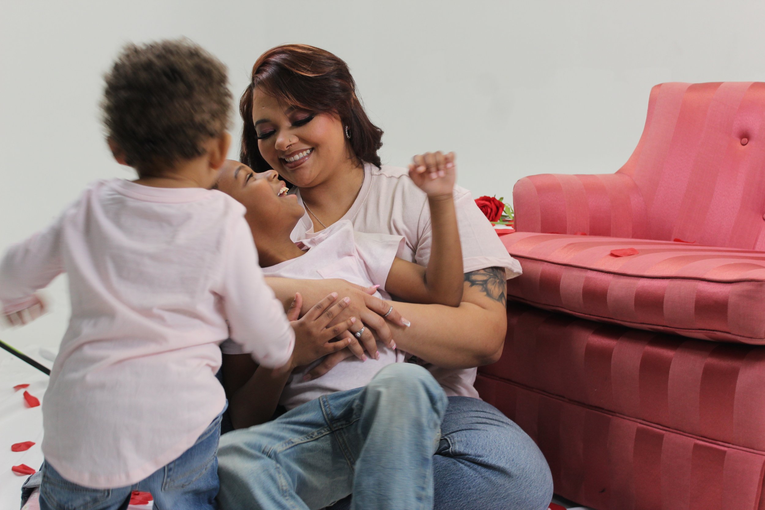 A woman and two children playing and laughing on the floor, with the woman sitting next to a pink sofa.