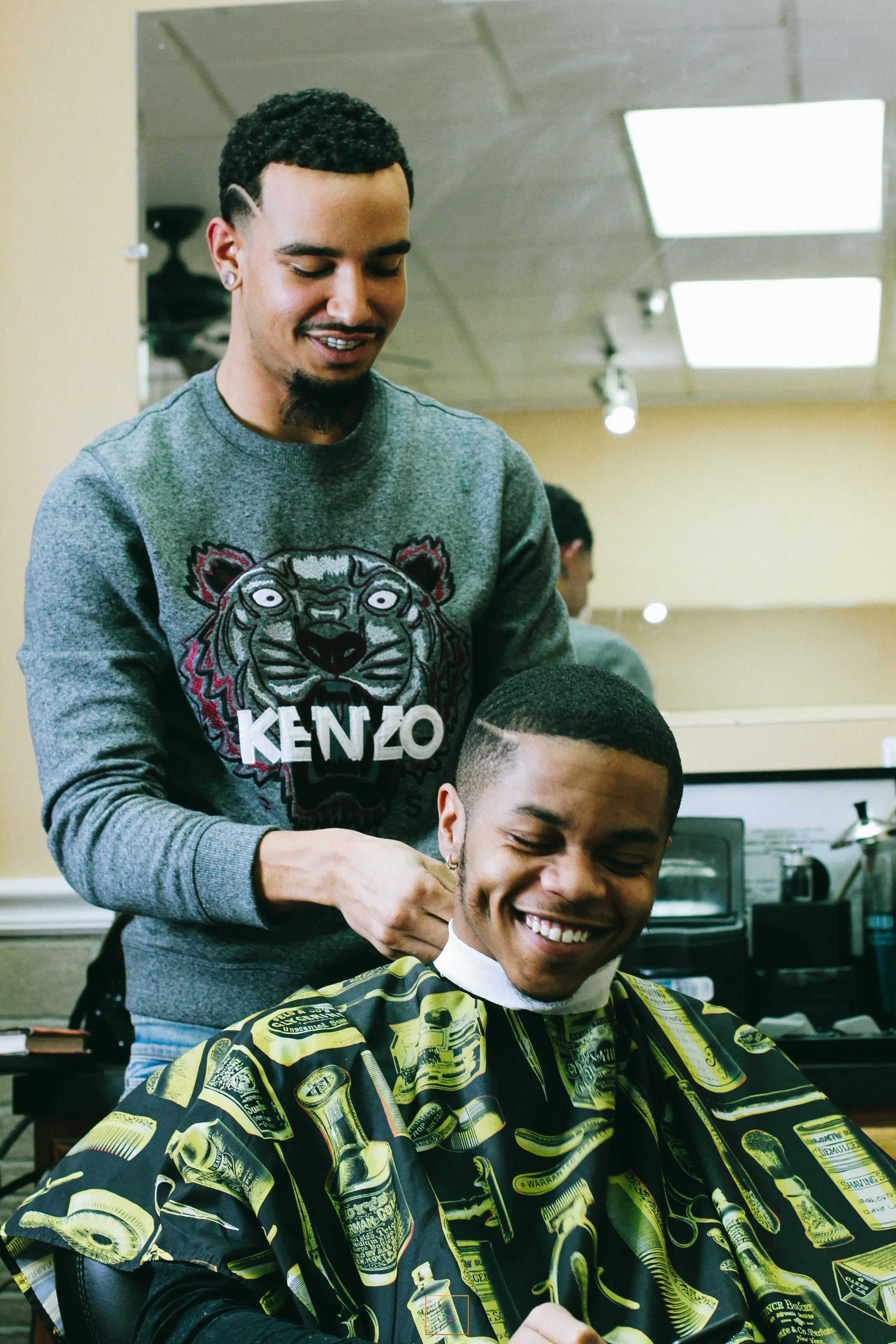 A young man with short hair and a goatee gets a haircut at a barbershop by a barber wearing a gray Kenzo sweatshirt with a lion design. The young man is smiling, wearing a barber cape with a print of bottles and cans.