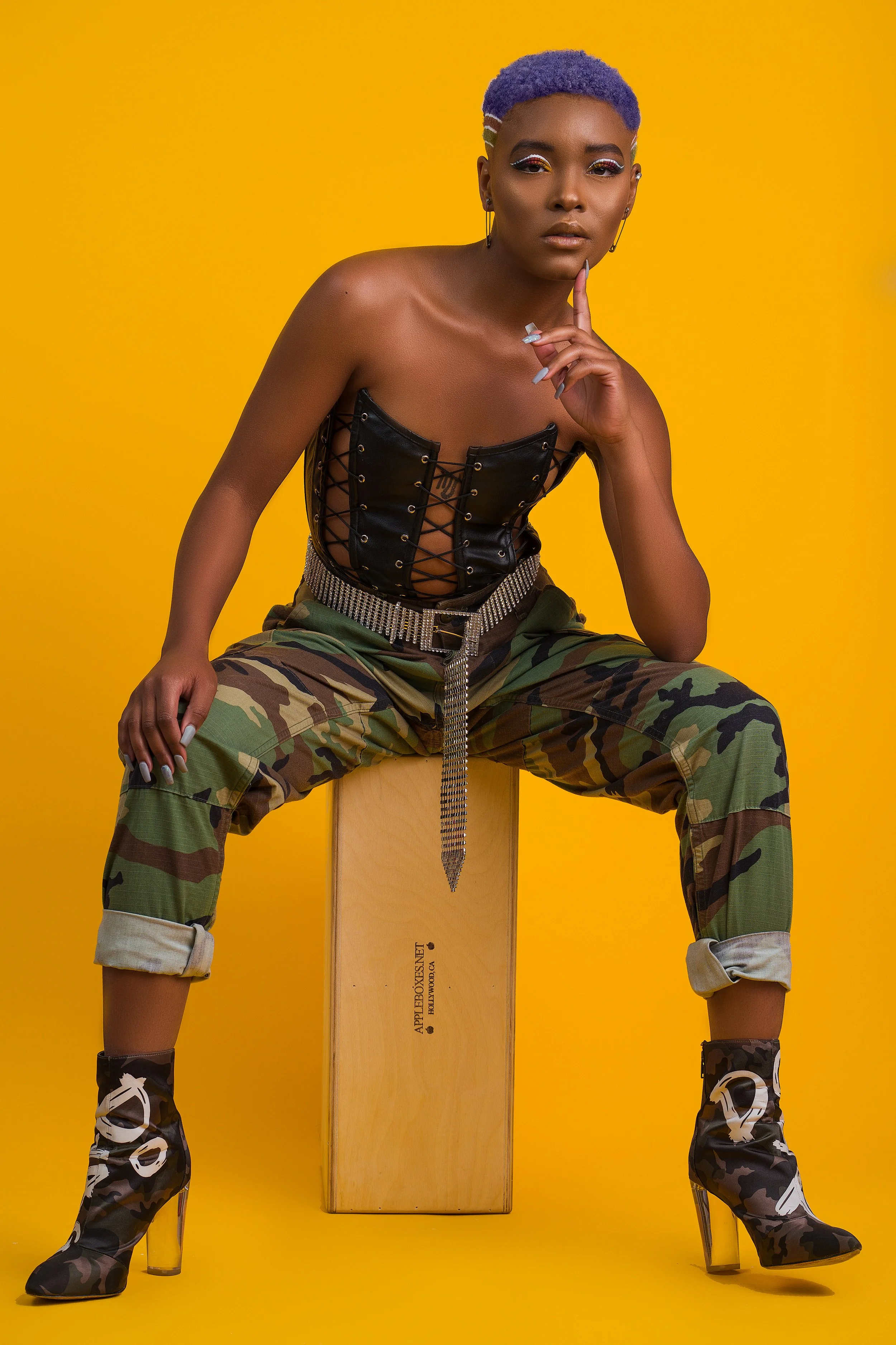 A woman with short purple hair sits on a wooden box against a yellow background. She is wearing a black leather corset top, camouflage pants with rolled-up cuffs, and high-heeled camouflage boots. She has eye makeup with glitter, earrings, and a beje