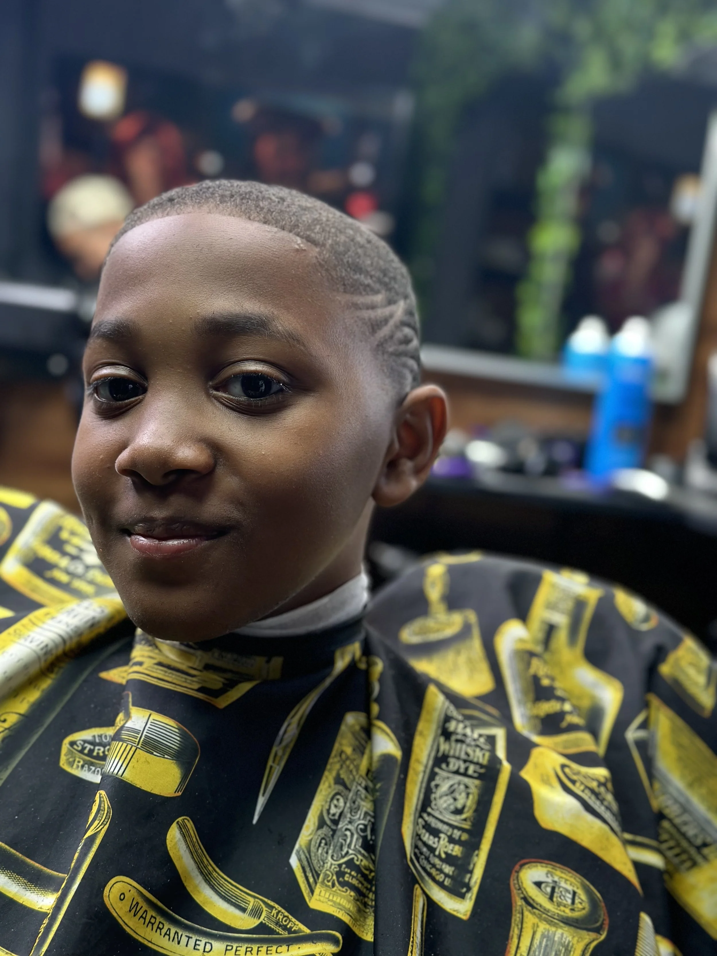 A young boy with freshly styled hair sitting in a barber's chair, wearing a black cape with yellow and white barber-themed graphics, in a barbershop with blurred mirrors and hair products in the background.
