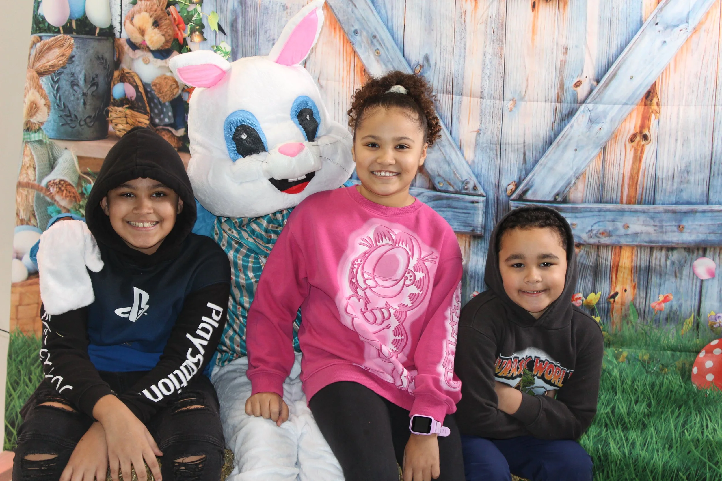 Three children sitting on a bench with a person dressed as a white rabbit in the background. The children are smiling, two boys and a girl, and are wearing casual hoodies and a pink sweater. The background features a painted farm scene with a wooden 