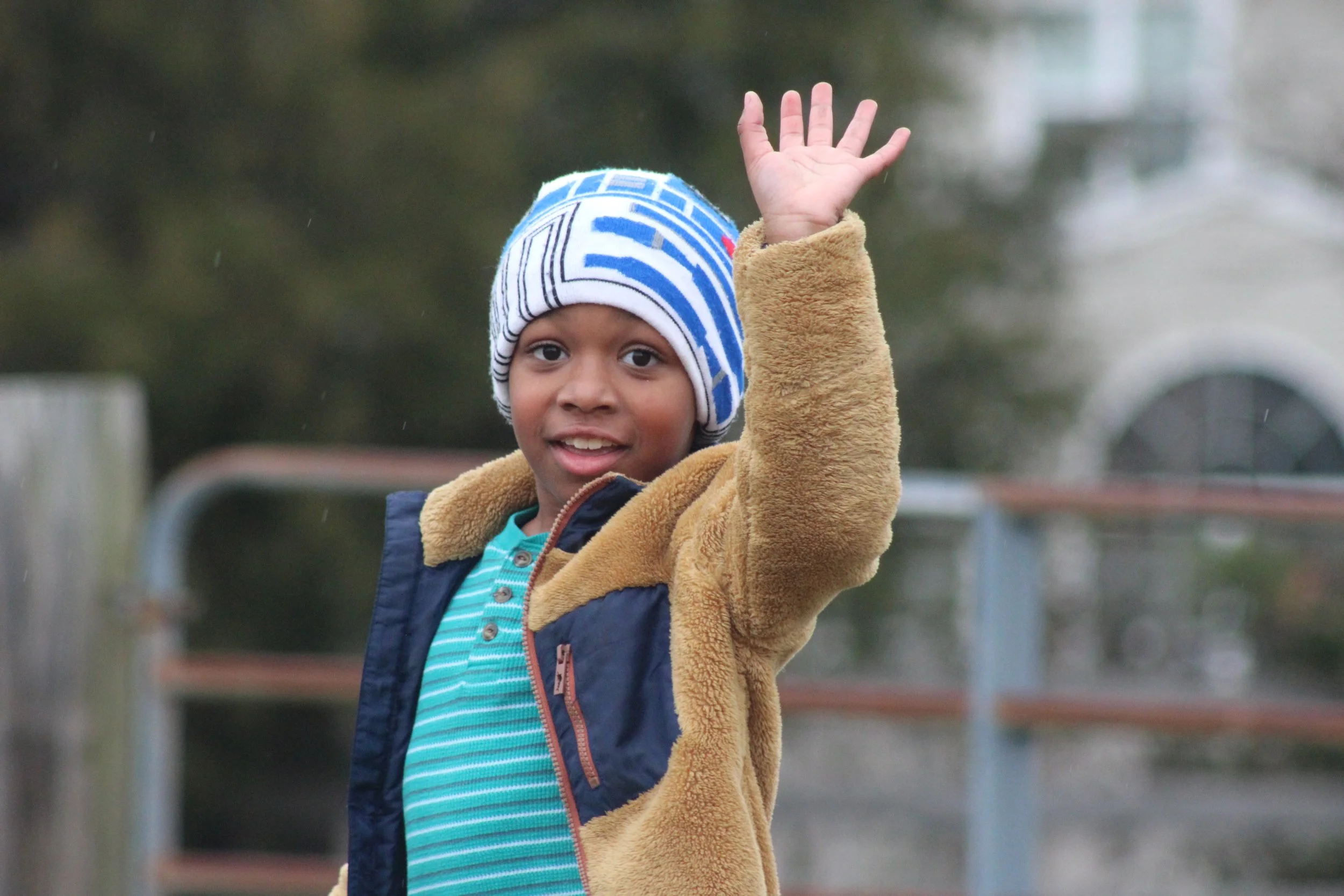 A young boy wearing a blue and white striped knit cap, a blue striped shirt, and a tan fleece jacket raising his hand outdoors.