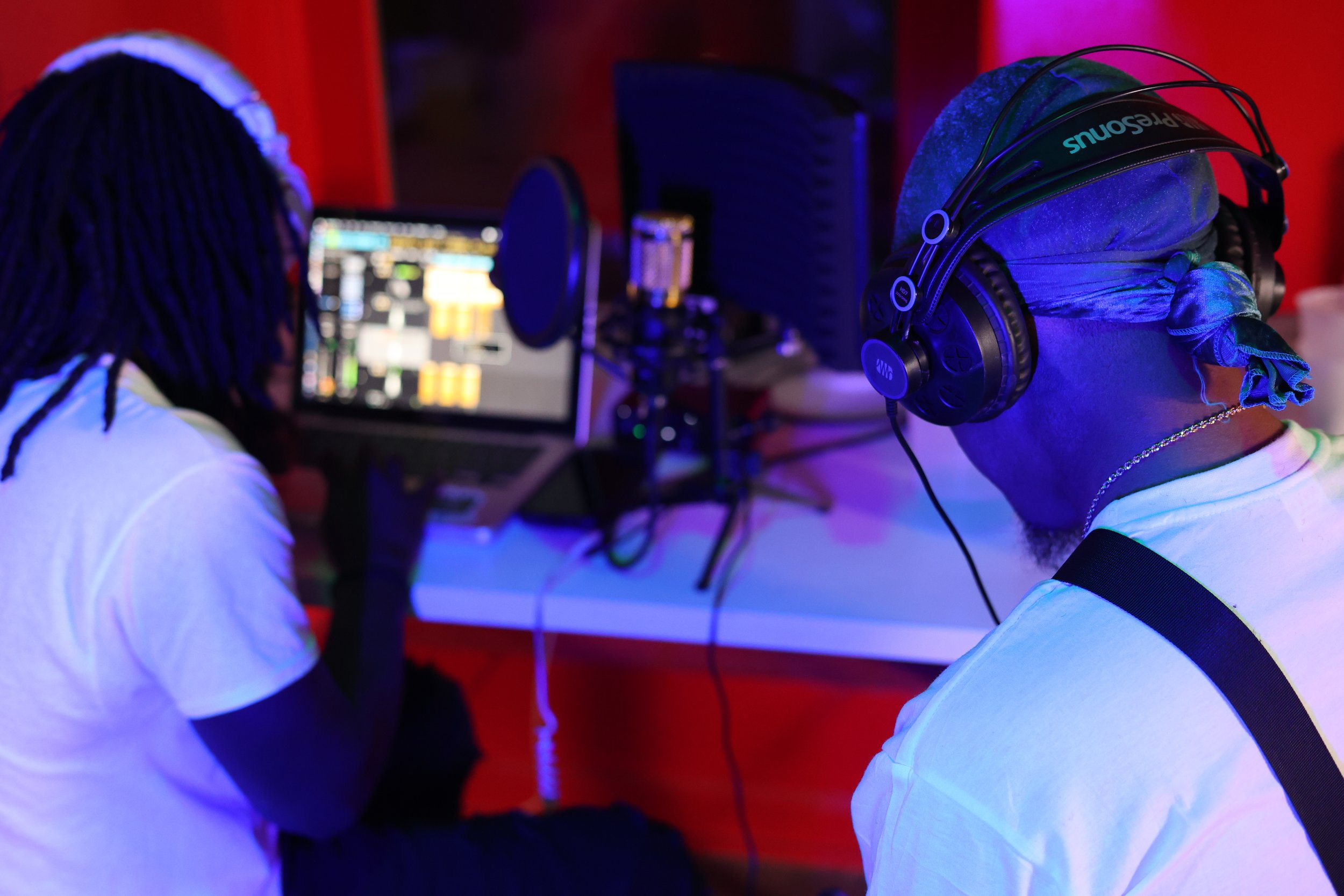 Two men in a recording studio working on music production, one with headphones and a bandana, and the other with dreadlocks, looking at a mixing console and computer screens.