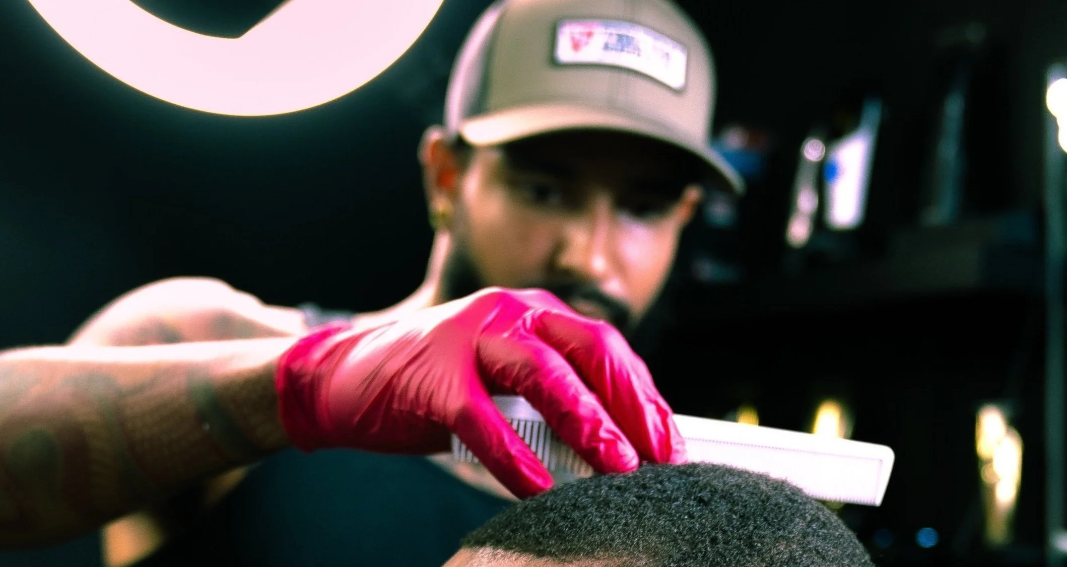 A person wearing a beige cap and pink gloves is using a brush to groom a person's short, curly hair in a salon or barber shop.