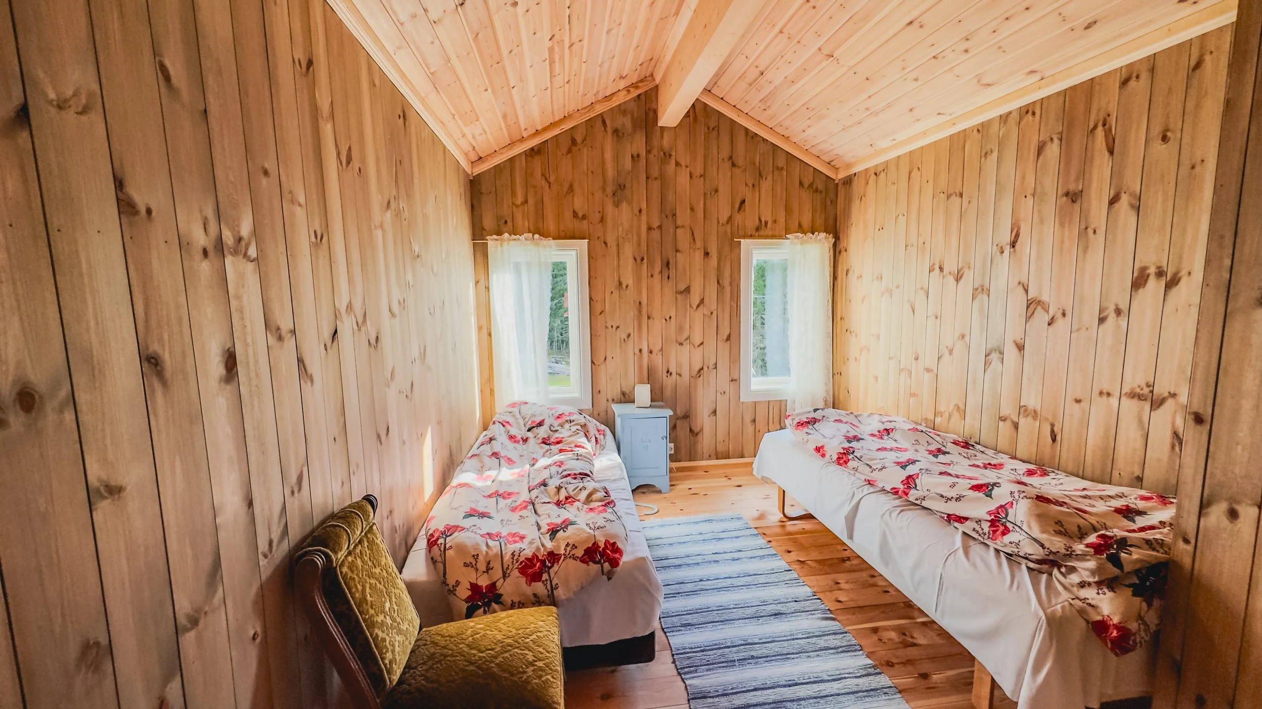 To enkeltsenger i et rom med trepanel i treverk og to vinduer med hvite gardiner.