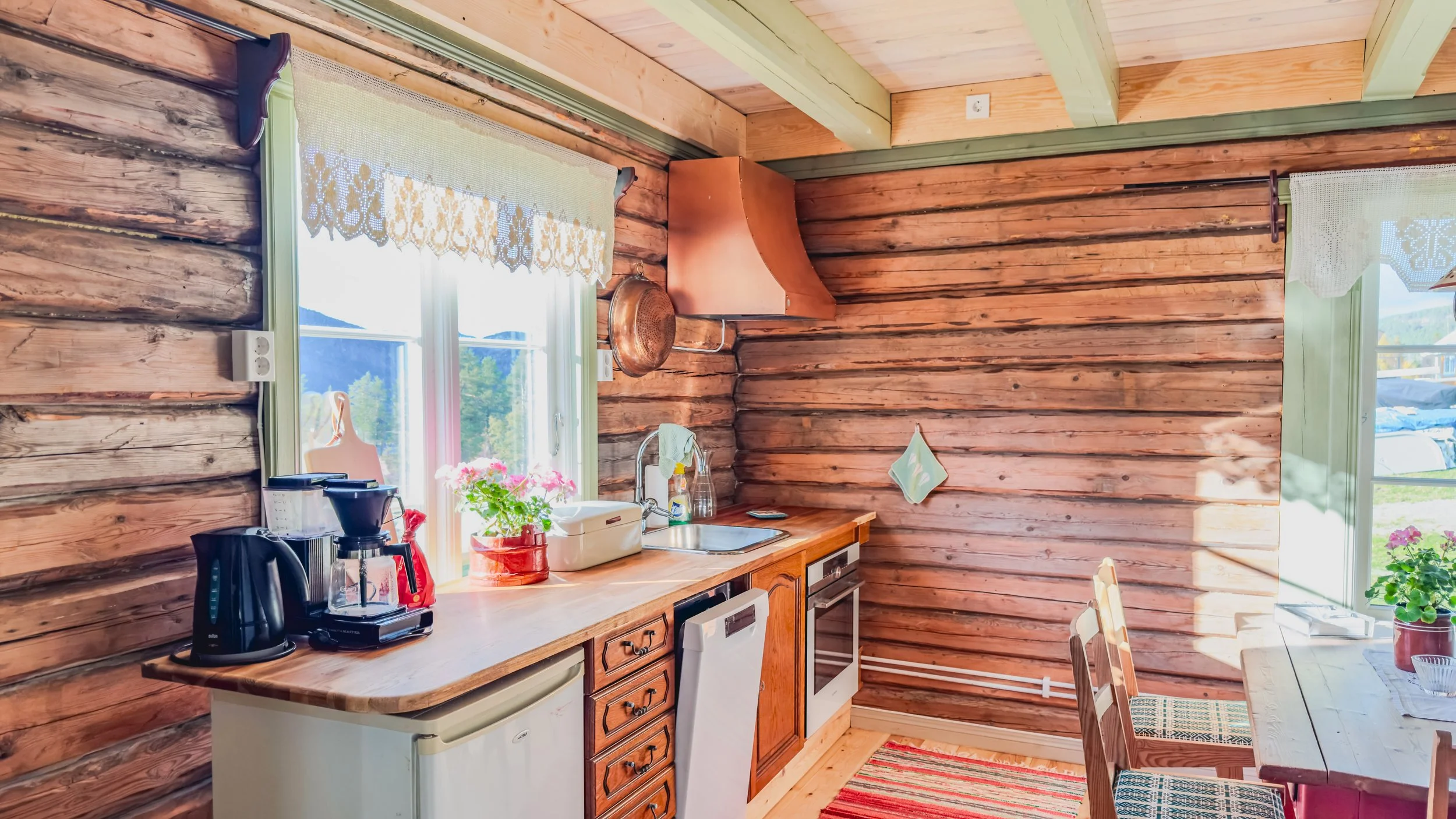 Kjøkken med rustikke tremøbler, vinduer med gardiner og blomster på bordet, utsikt mot utendørsområde med utendørsmøbler.