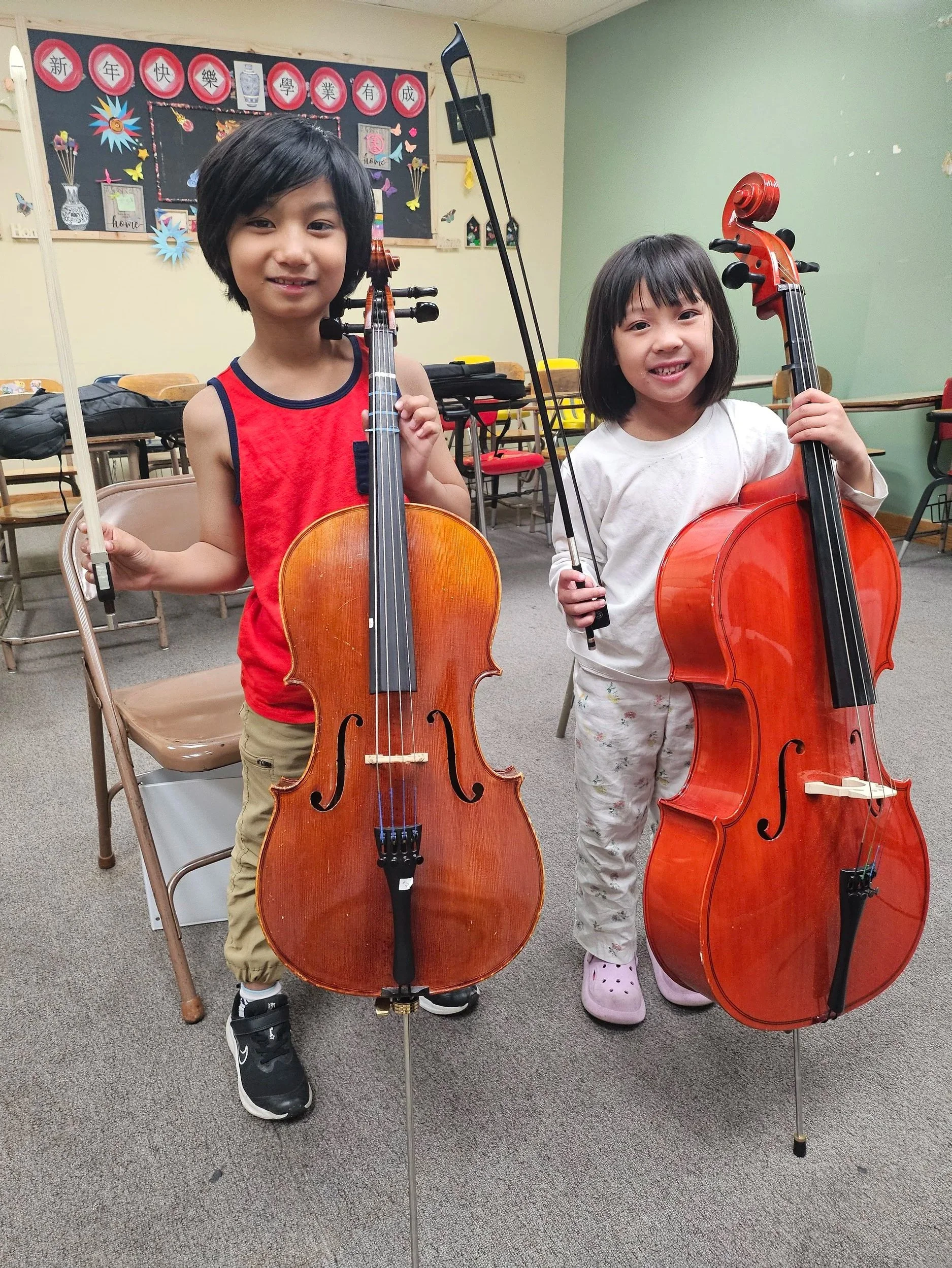 Two young girls standing in a classroom, holding violins and bows, smiling at the camera.