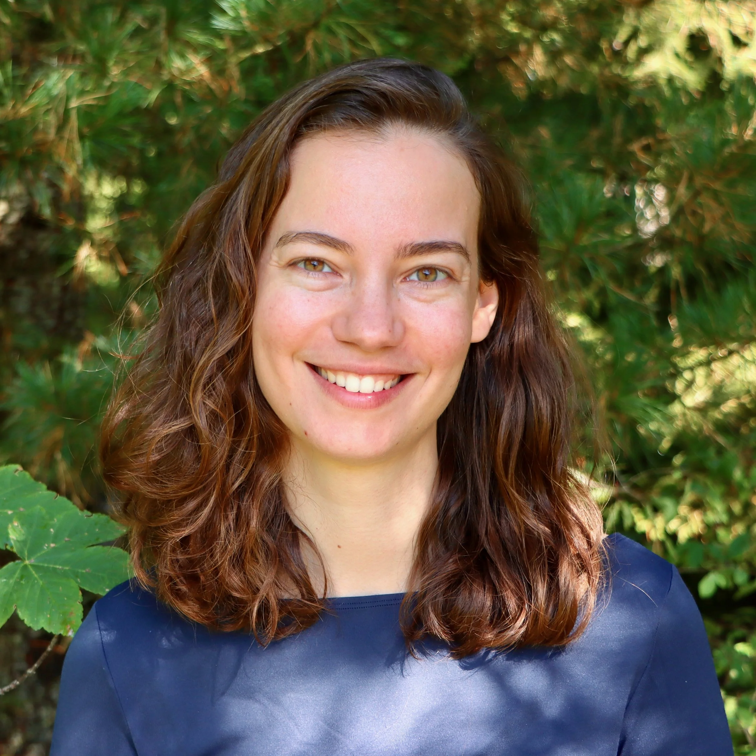 Portrait of a young woman with brown, wavy hair, smiling, wearing a navy blue top, outdoors with green foliage in the background.