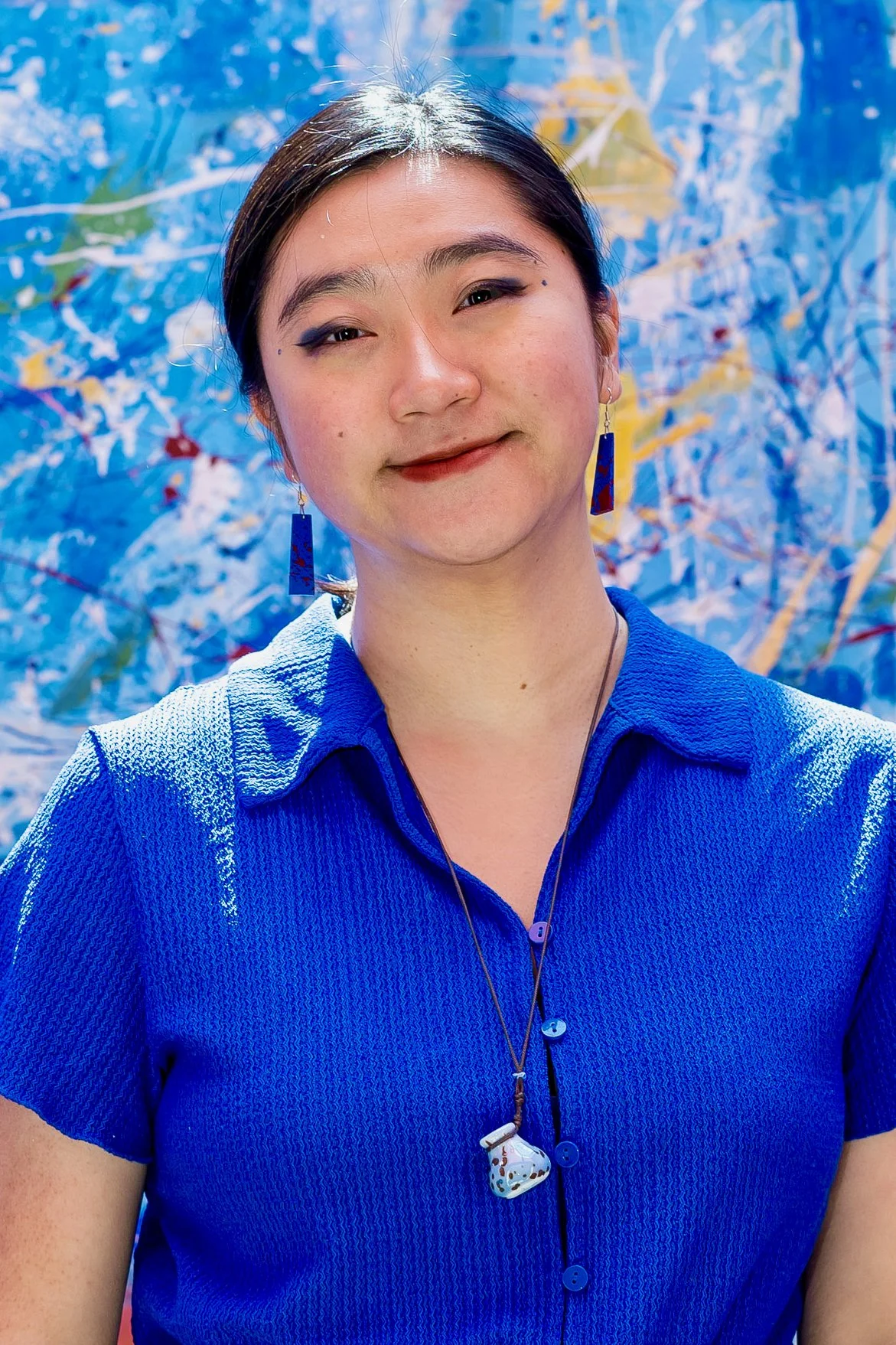 A woman with dark hair wearing a blue shirt, colorful earrings, and a necklace with a spoon pendant, smiling and winking against a bright, abstract blue background.
