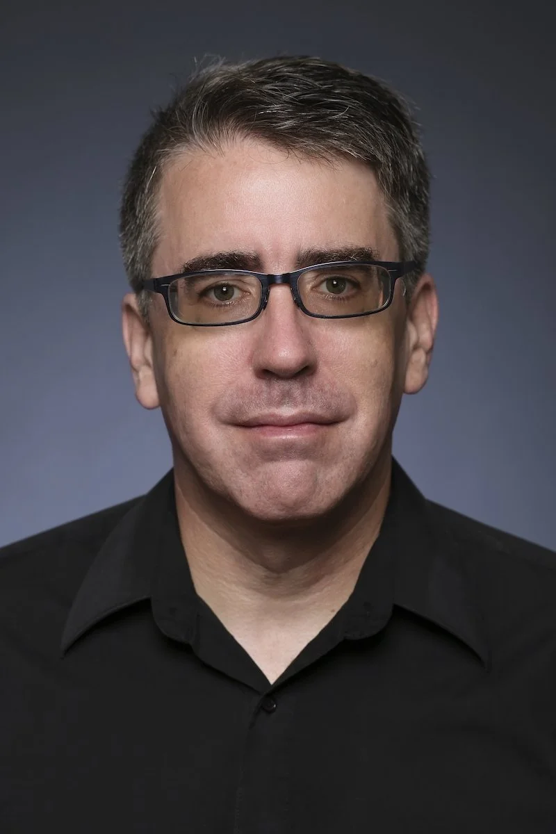 A man with short brown hair, wearing black glasses and a black collared shirt, against a plain dark background.