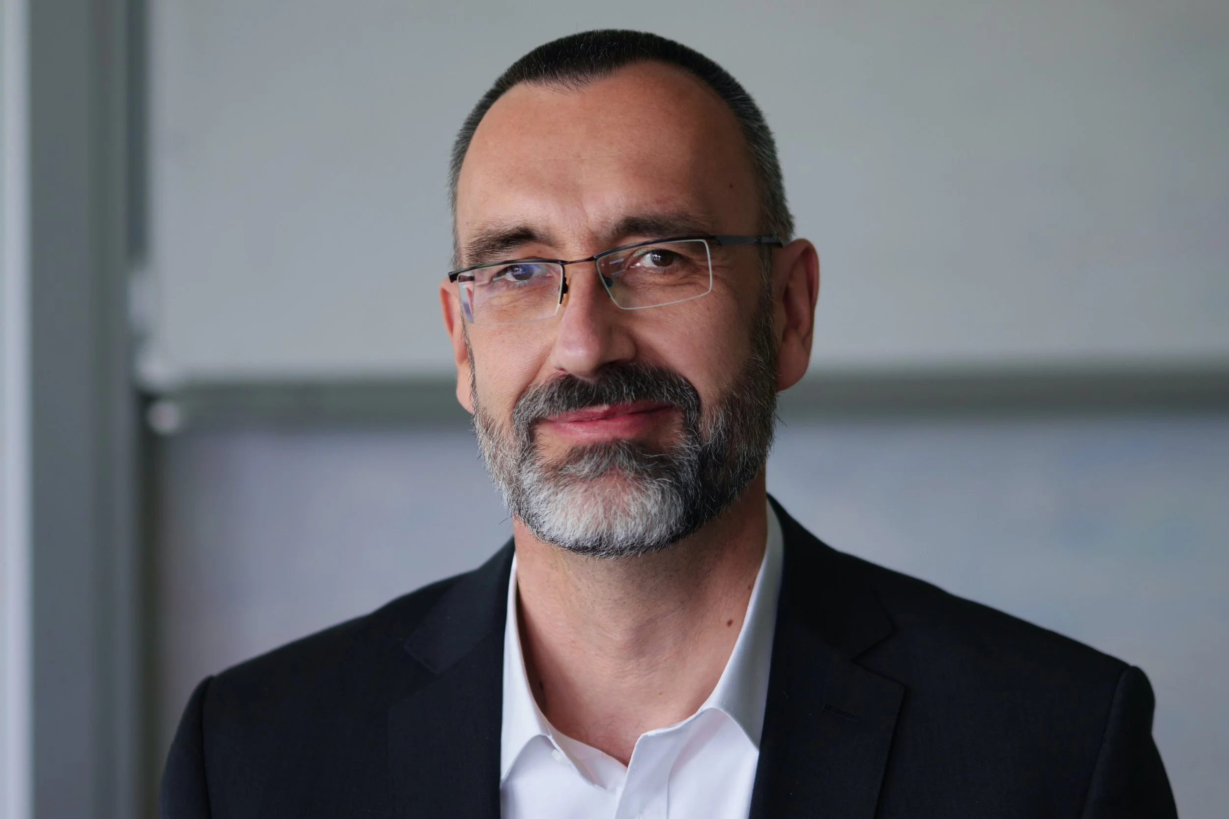 A middle-aged man with glasses, a gray beard, wearing a suit and white shirt, looking at the camera with a slight smile, in an office setting.