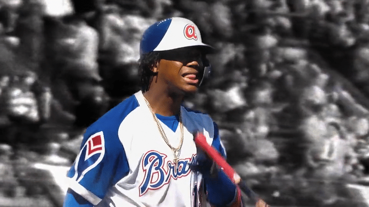 A person wearing a baseball helmet and a jersey with the word 'Braves' on it, standing outdoors with rocks in the background.