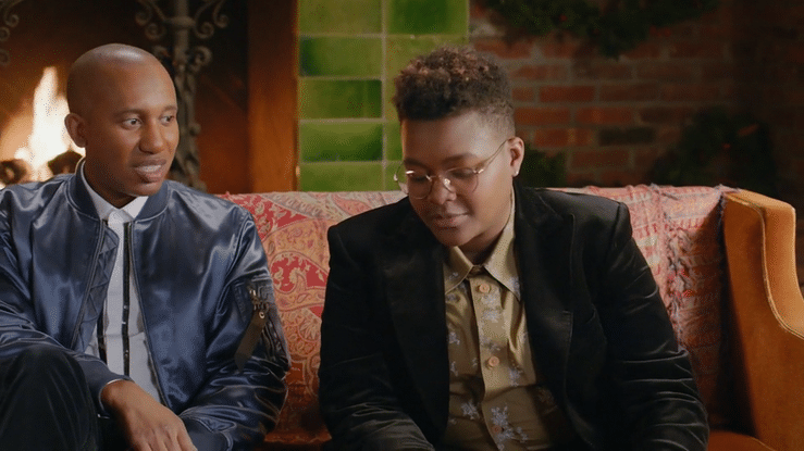 Two young men sitting on a patterned sofa near a fireplace and a green window, engaged in conversation.