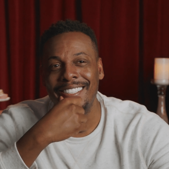 A smiling man with short curly hair resting his chin on his hand in front of a red curtain, with a lit lamp on his right.