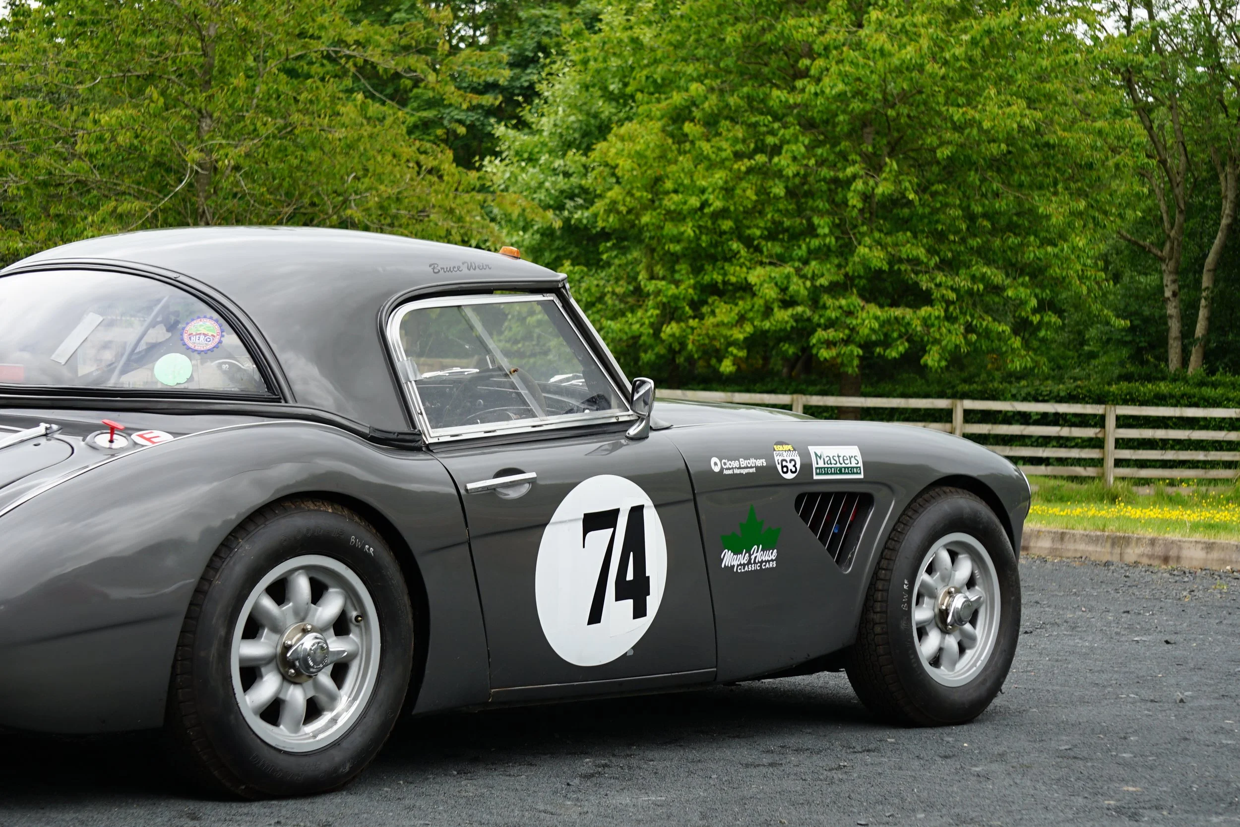 A classic gray racing car with the number 74 on the door, parked on a gravel surface, with green trees and a wooden fence in the background.