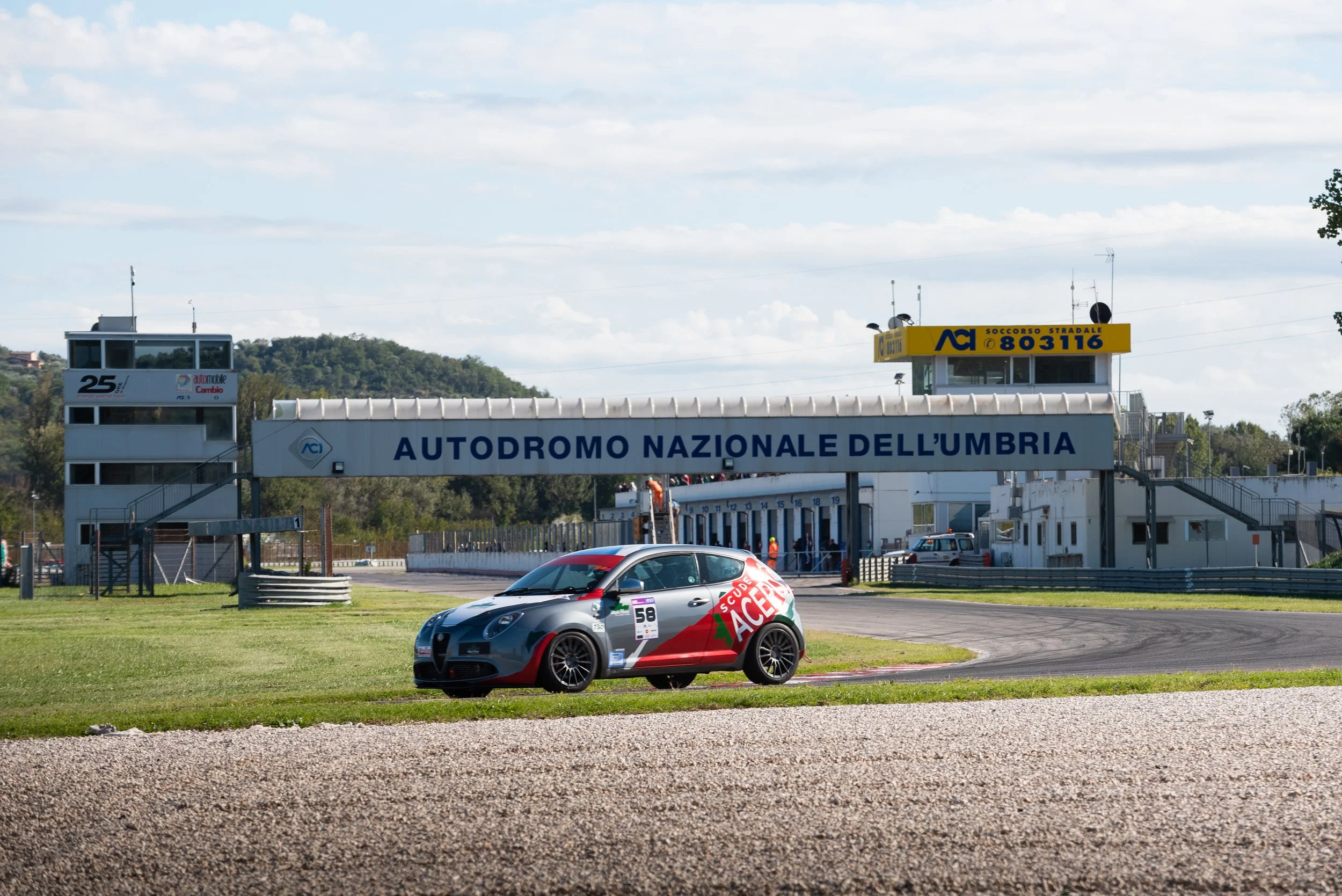 A race car with the number 58 on it parked on a track at Autodromo Nazionale dell'Umbria, with a grandstand and control tower in the background.
