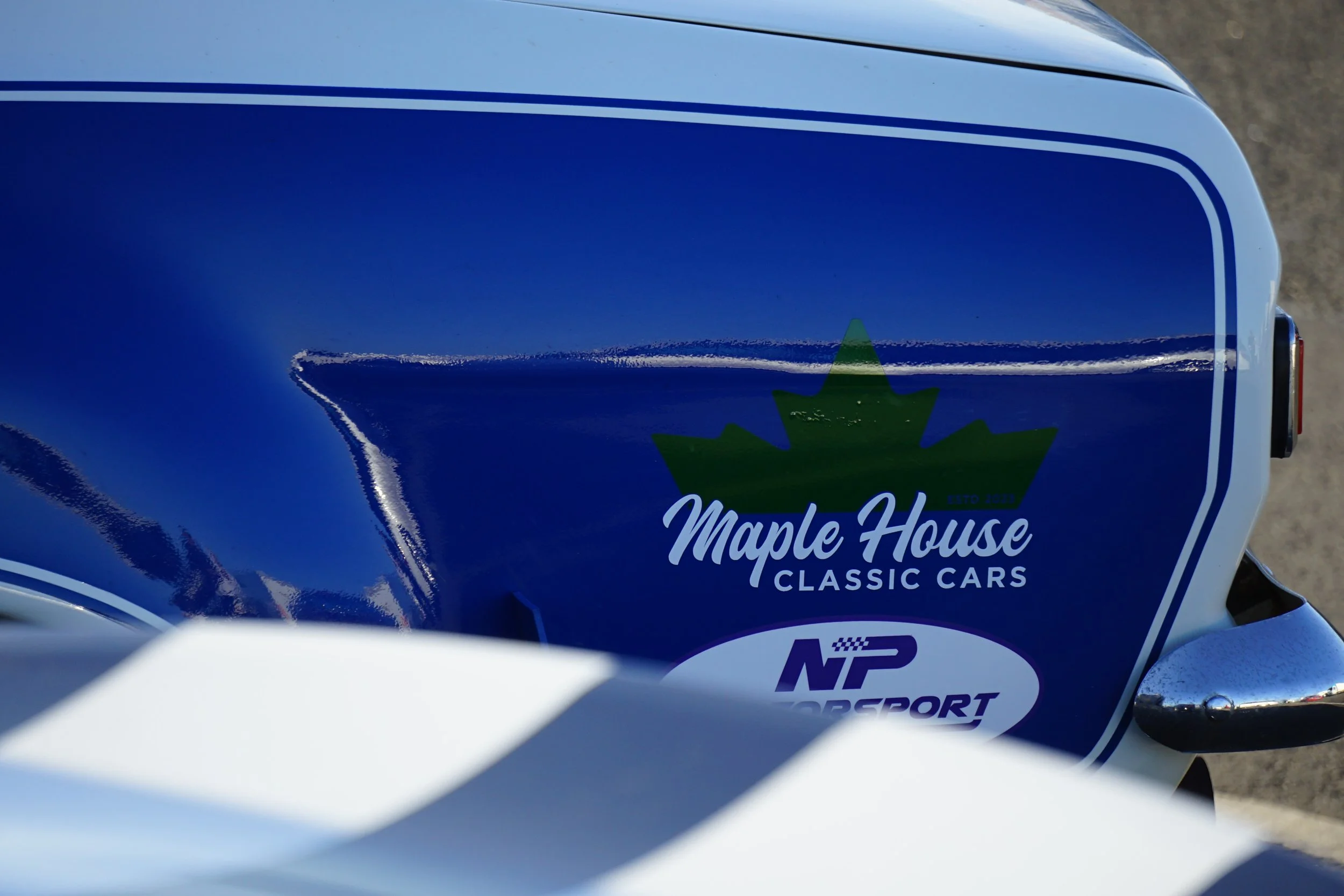 Close-up of a blue car with Maple House Classic Cars and NP Motorsport stickers on the side.
