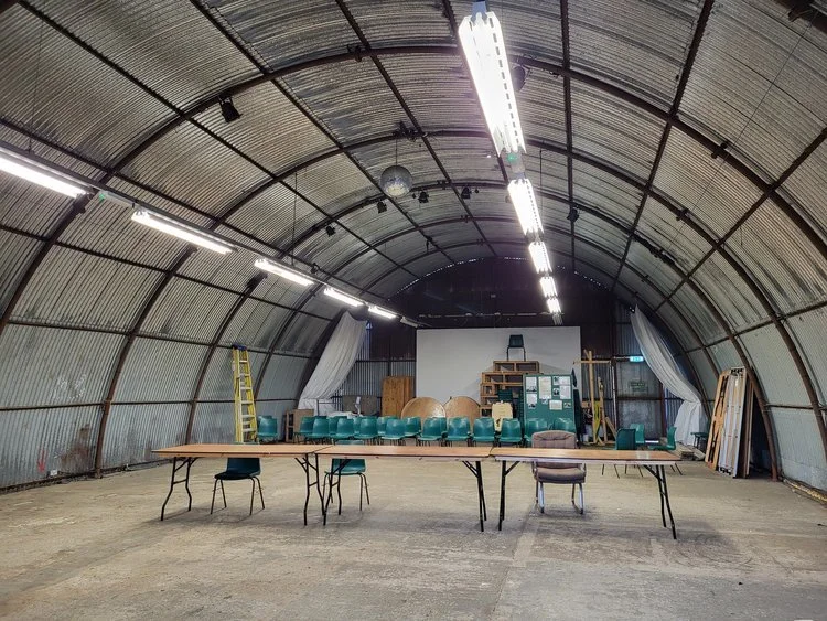 Interior of a large, industrial-style arched building with exposed metal walls, featuring long tables and several chairs. The space is equipped with fluorescent lighting, a ladder, stacked seating, and various equipment and materials along the back wall.