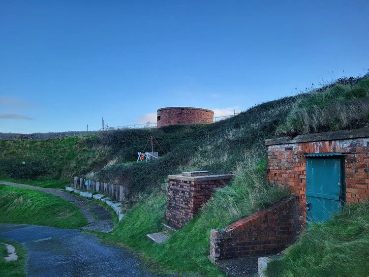 Landscape with brick structures, grassy hills, and pathway under clear blue sky.