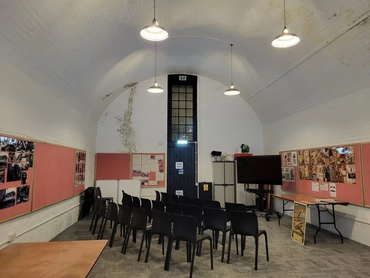 Room with arched ceiling, rows of black chairs, bulletin boards with photos, and a TV screen.