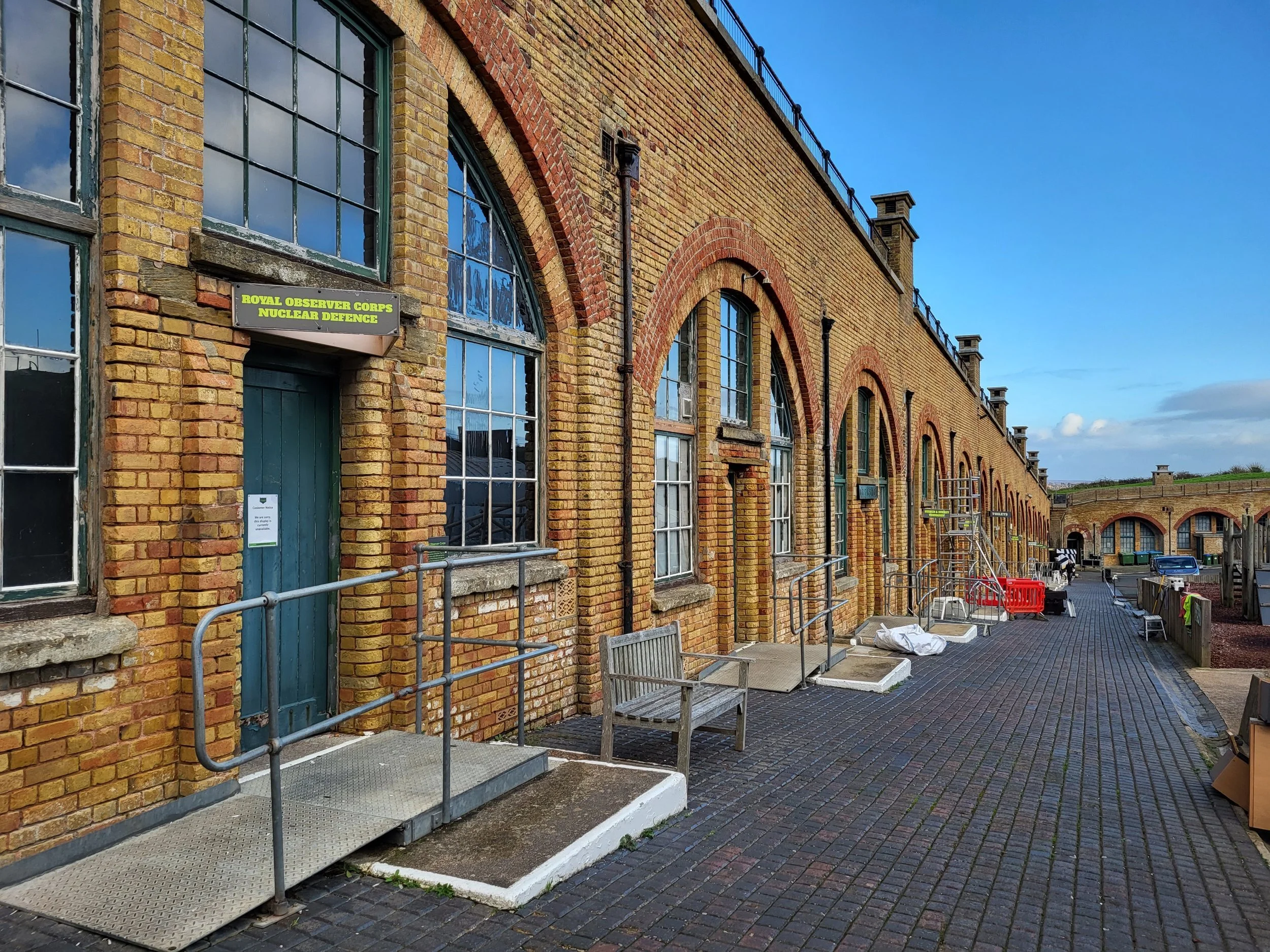 A long brick building with arched windows and a ramp outside. A sign on the building reads "Royal Observer Corps Nuclear Defence." The area has benches, a ladder, and construction materials. A blue sky is visible above.