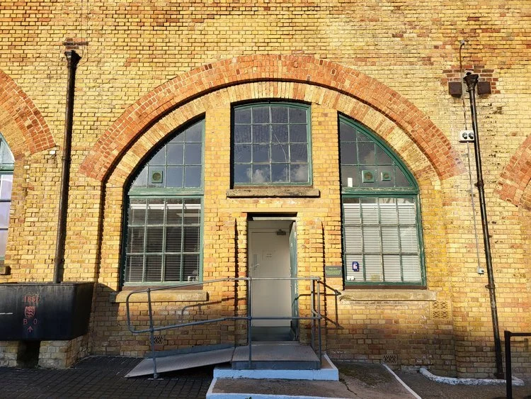 Brick building with arched windows and central door, featuring a wheelchair ramp and railings.