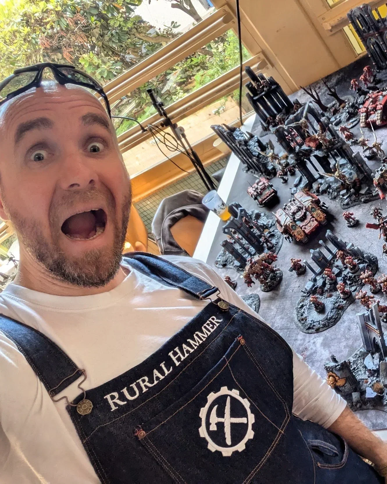 Un homme souriant avec des lunettes sur la tête portant un tablier avec l'inscription 'Rural Hammer' pose devant un plateau de figurines de jeux de guerre peintes. Il semble fier de ses figurines. La pièce est éclairée par la lumière naturelle venant d'une fenêtre en arrière-plan.