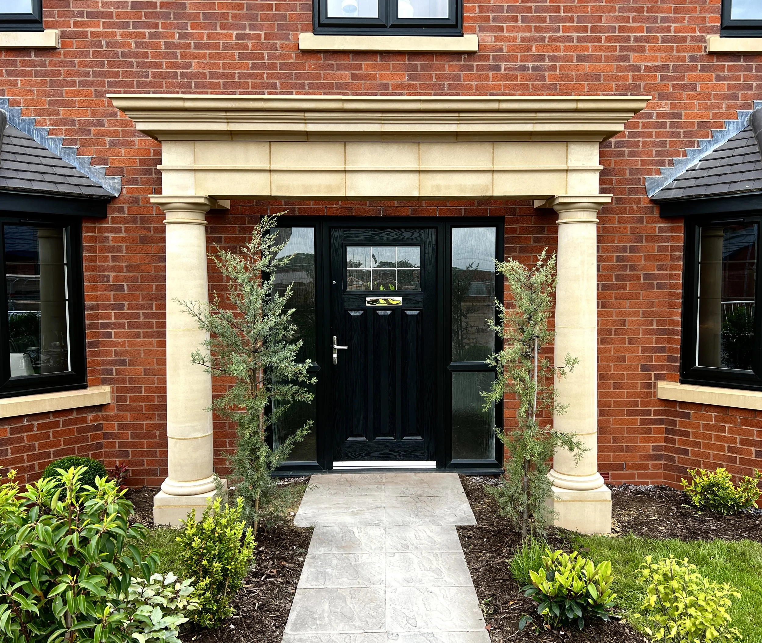 Front entrance of a brick house with a black door, flanked by two classical columns with plants on either side, and a pathway leading to the door.