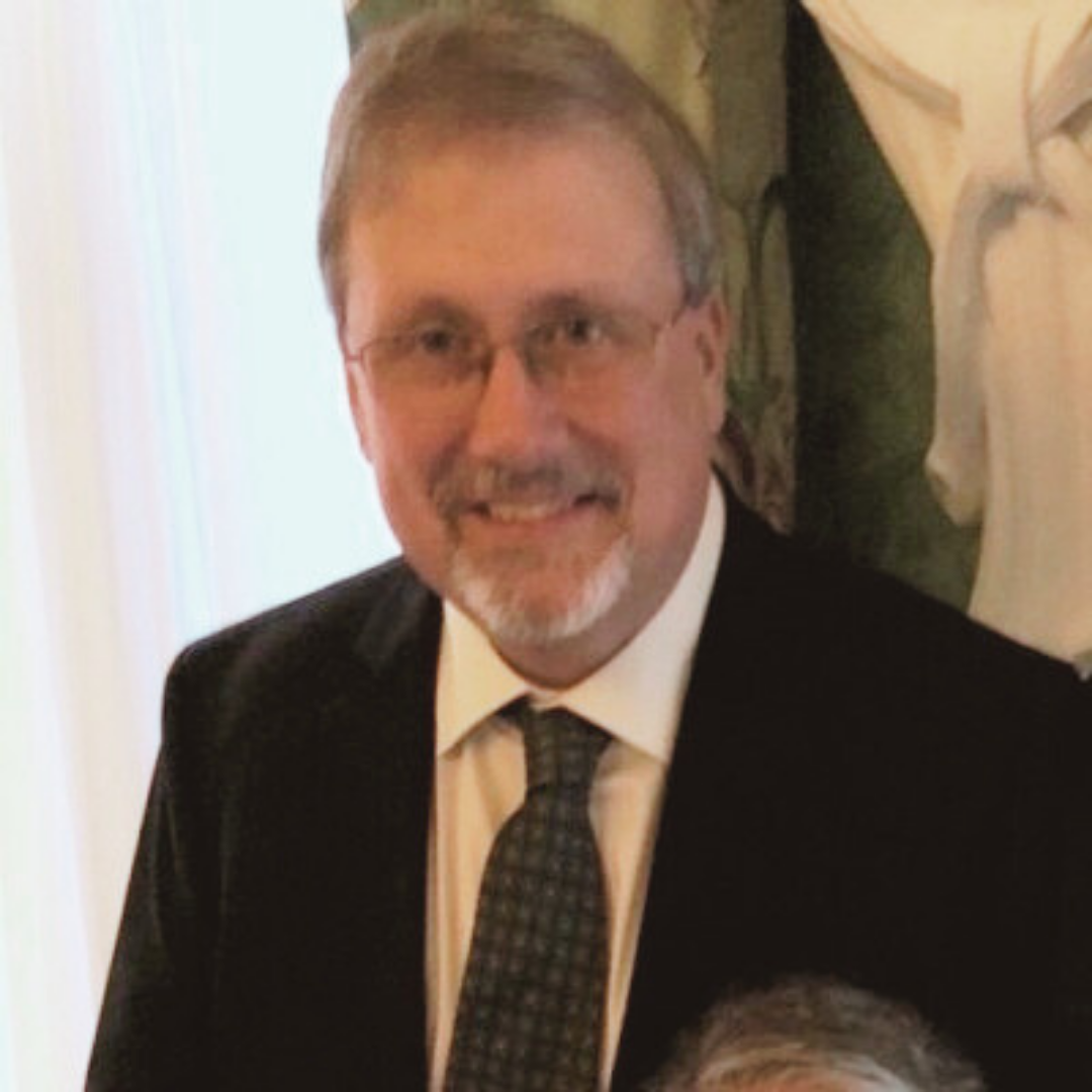 A man with glasses, gray hair, and a beard smiling, wearing a suit and tie, in front of a white wall and a decorative curtain - representing a happy client.