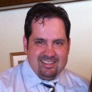 A smiling man in a dress shirt and tie, indoors with a framed picture in the background - representing a happy client.