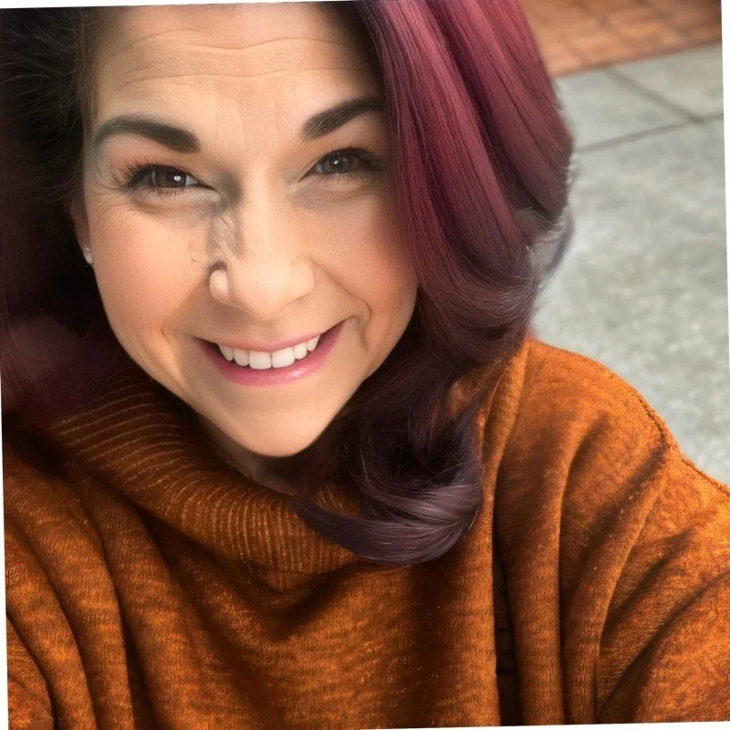 Close-up of a smiling woman with dark blonde to red hair, wearing a brown turtleneck sweater, sitting outdoors on a concrete surface.