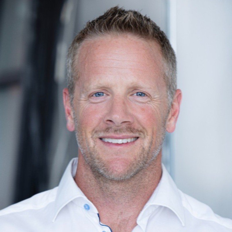 Smiling man with short, light brown hair and blue eyes, wearing a white dress shirt, in an indoor setting with blurred background.