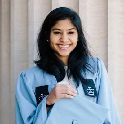Young woman in a light blue graduation gown smiling, standing in front of beige curtains  - representing a happy client.