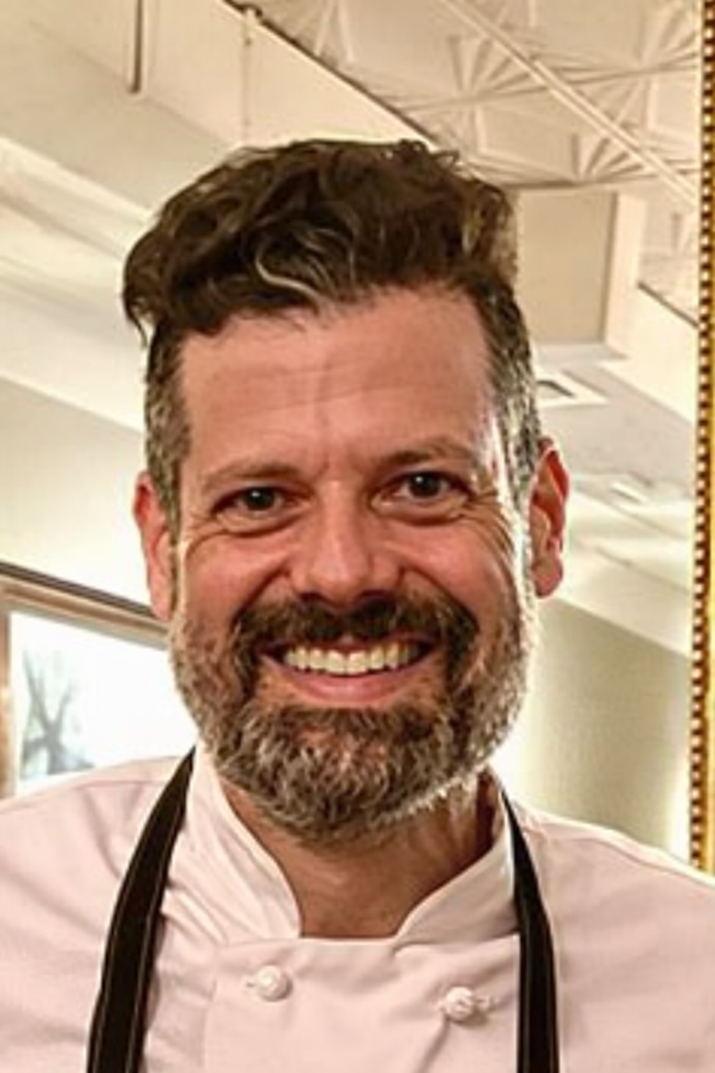 A smiling man with curly brown hair, a beard, and mustache, wearing a chef's coat and black apron, indoors with a ceiling and window in the background.