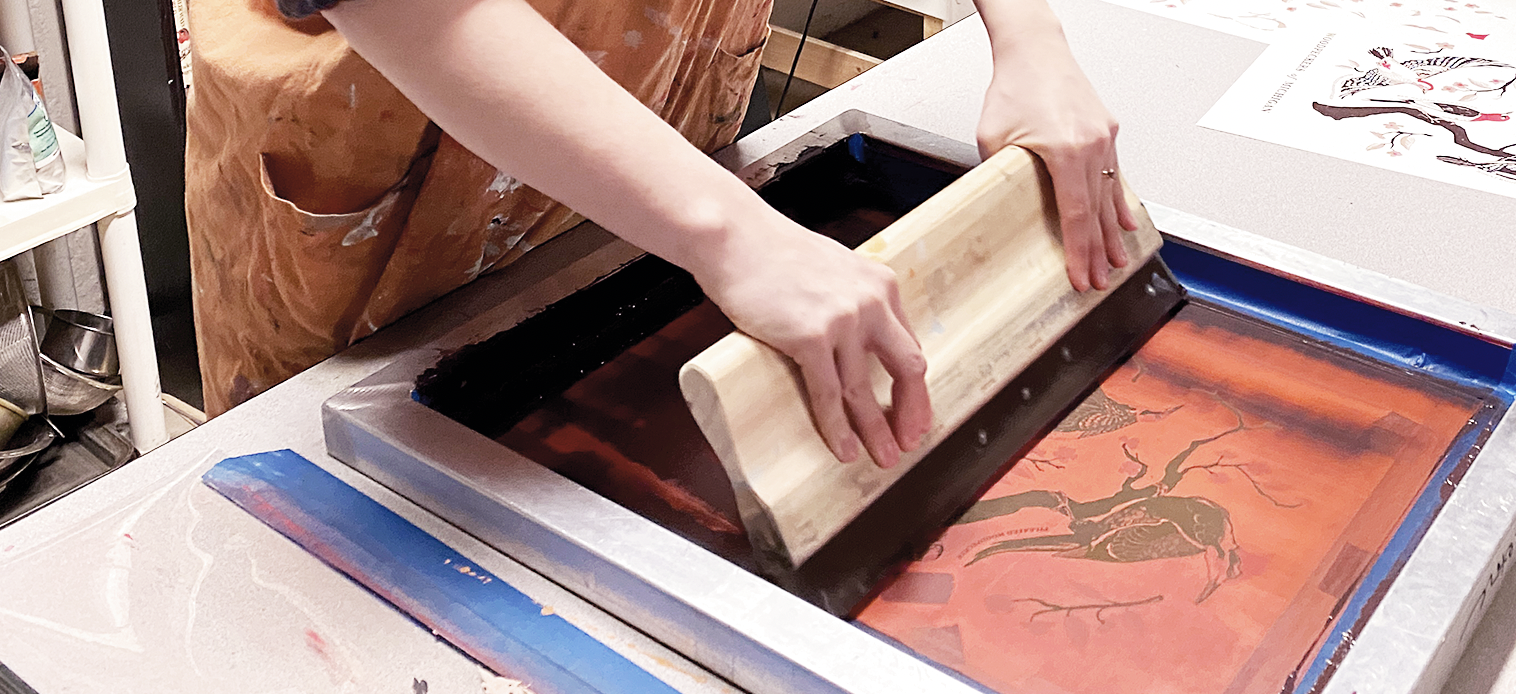 Person using a squeegee to press paper on a wooden frame during printmaking in a studio.