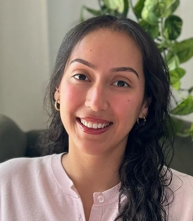 A woman with long dark curly hair, wearing a light pink button-up shirt, sitting on a gray chair in a room with white curtains and green plants.
