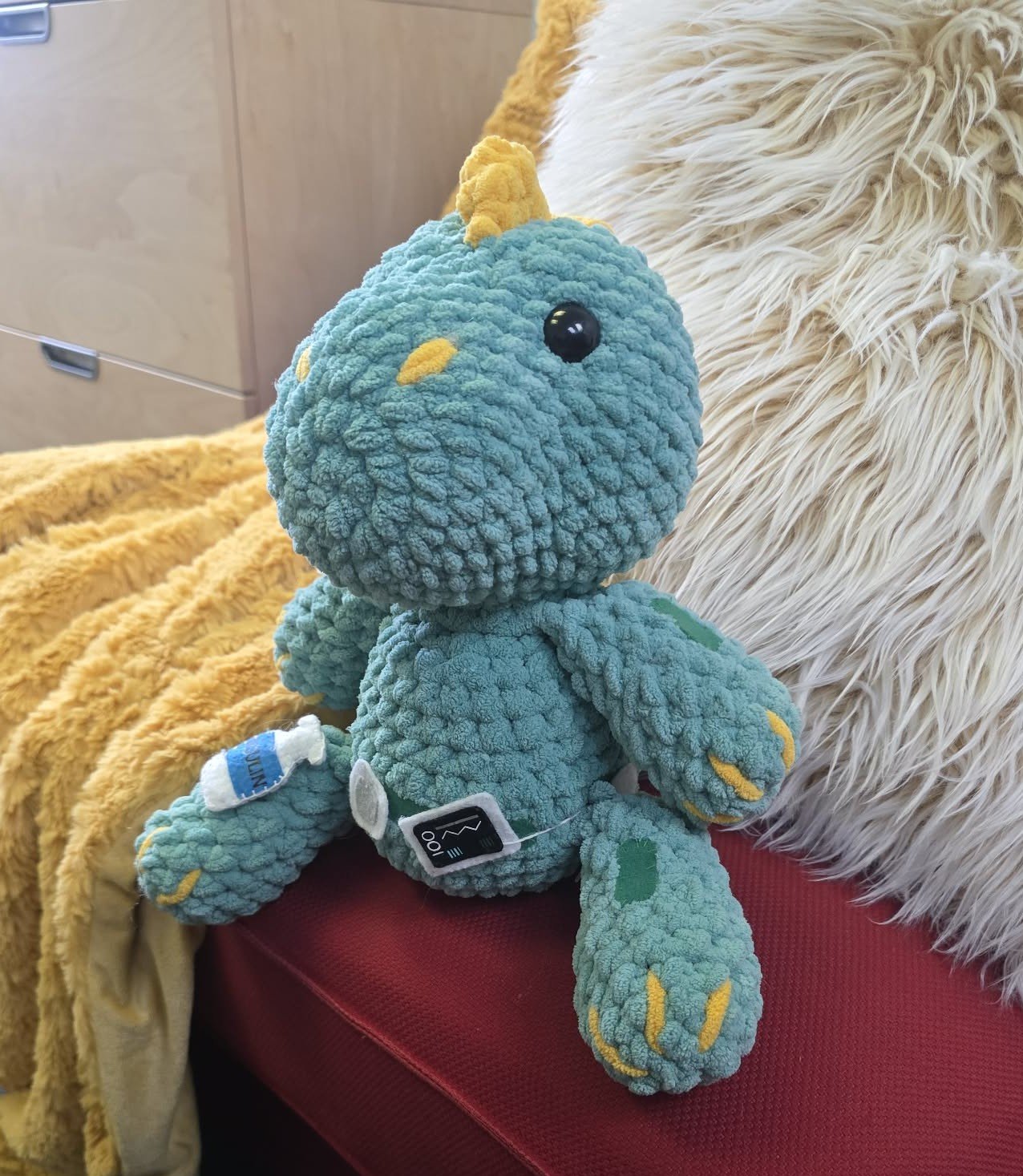 A plush toy dinosaur with blue fluffy texture, yellow accents, and a yellow horn, sitting on a red surface next to a cream-colored fluffy blanket and pillow.