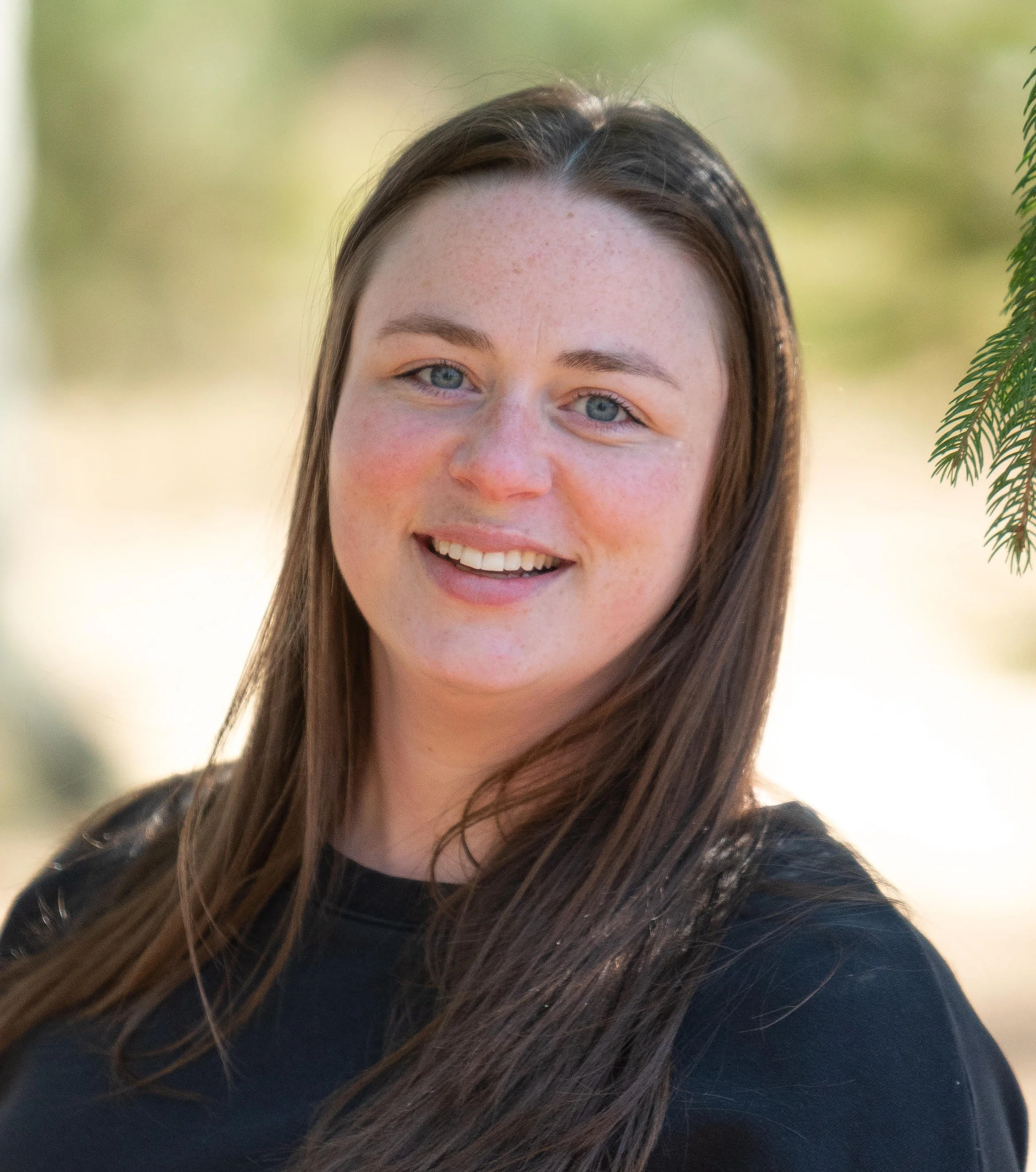 A young woman with long brown hair and blue eyes smiling outdoors with a blurred green background and hanging tree branches.