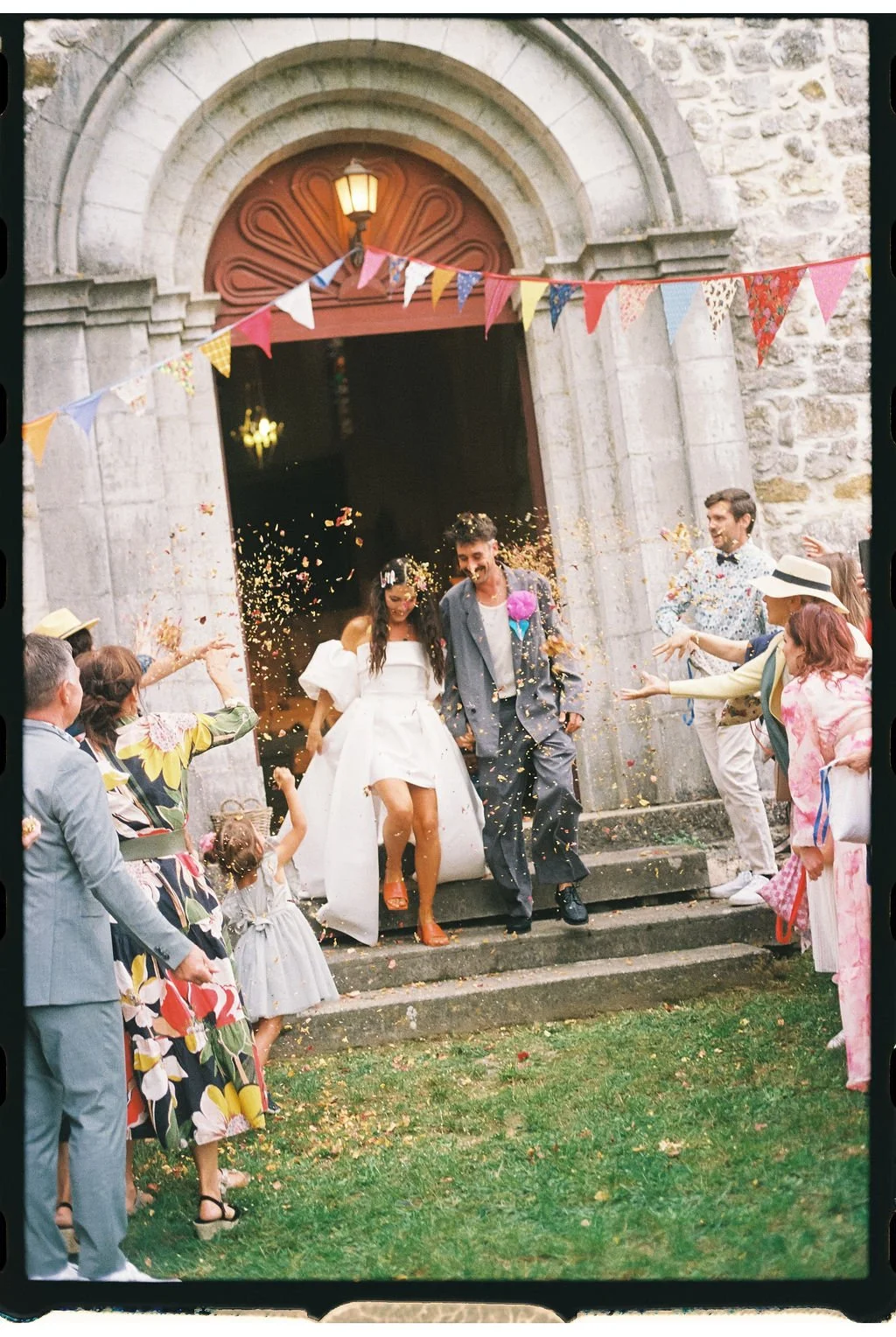 Jeunes mariés sortant d’une église en pierre sous des confettis, entourés d’invités et de fanions colorés.
