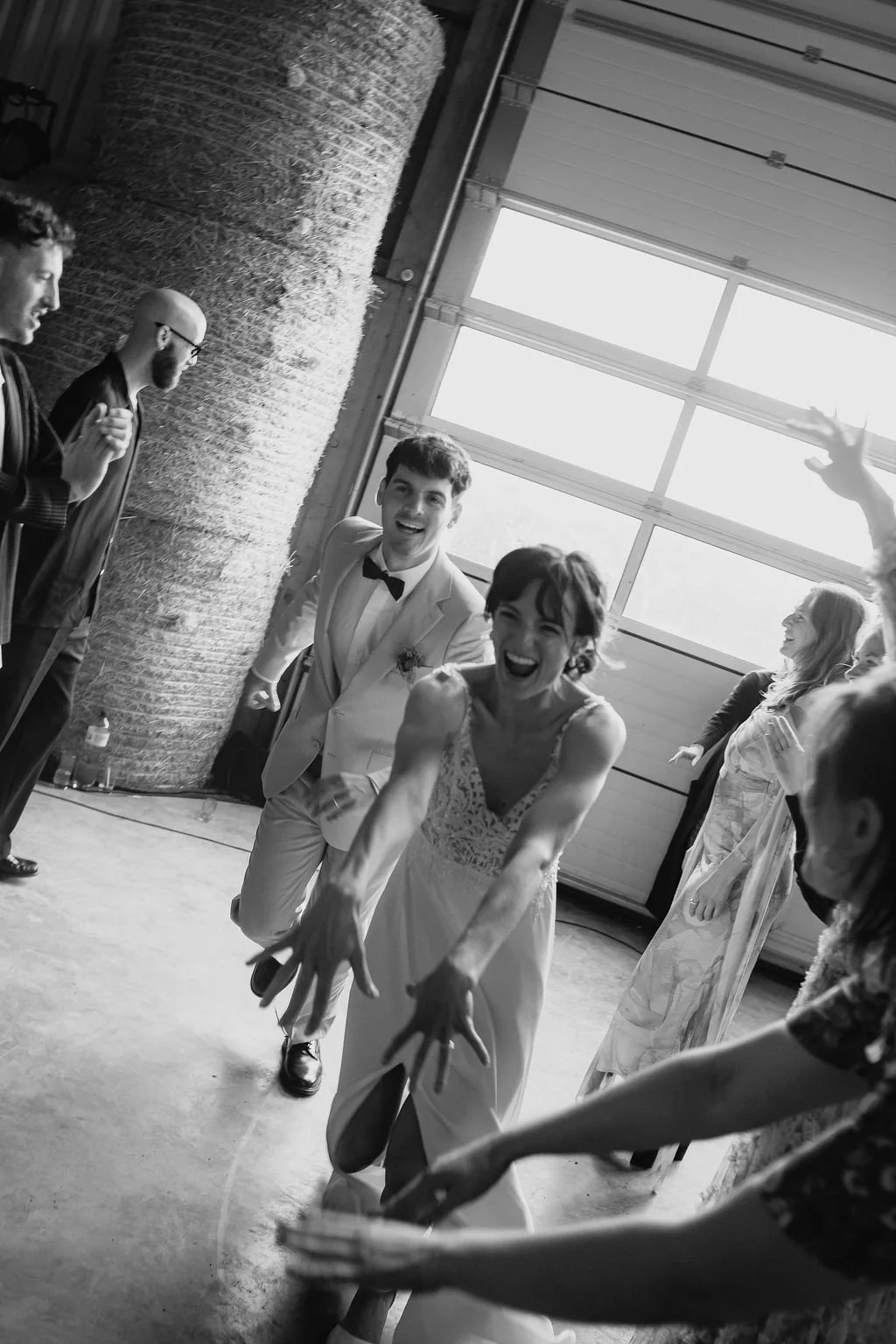 Une femme en robe de mariée souriante s'amuse lors d'une fête ou d'une cérémonie, entourée de plusieurs personnes, avec un homme en costume blanc et un nœud papillon qui apparaît également heureux.