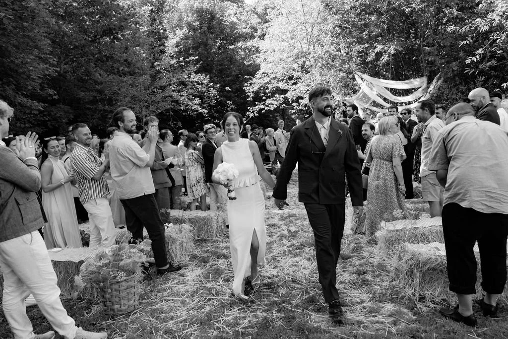 Jeunes mariés sortant d’une cérémonie de mariage en plein air, entourés d’invités dans un cadre forestier.