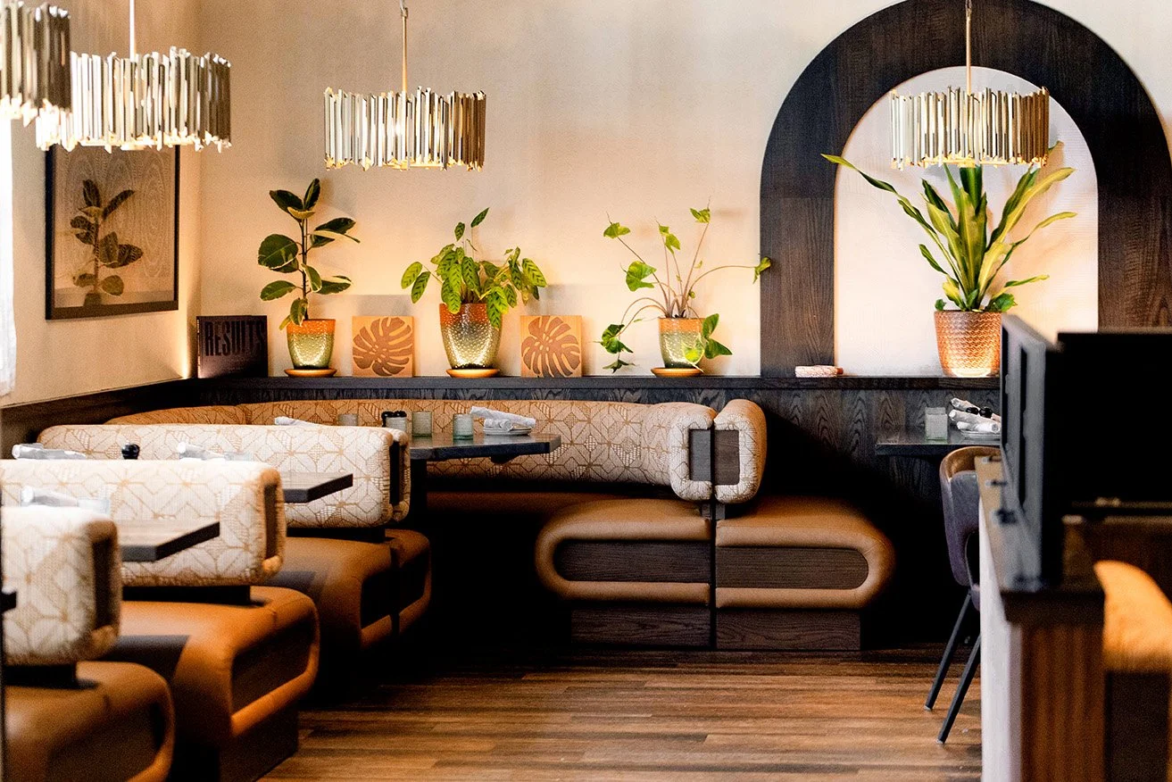A cozy restaurant interior with a dark wood and cream color scheme, featuring cushioned booths, potted plants on a ledge, framed botanical art, modern chandeliers, and a wooden floor.