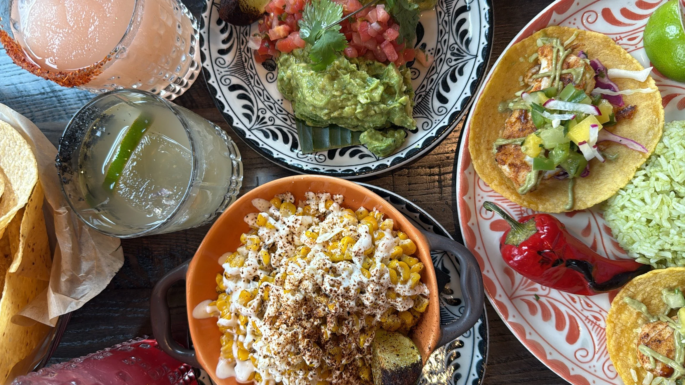 A table spread of Mexican dishes including guacamole with salsa, fresh lime, a corn salad, a spicy red pepper, and beverages like a pink cocktail and a light-colored drink with lime.